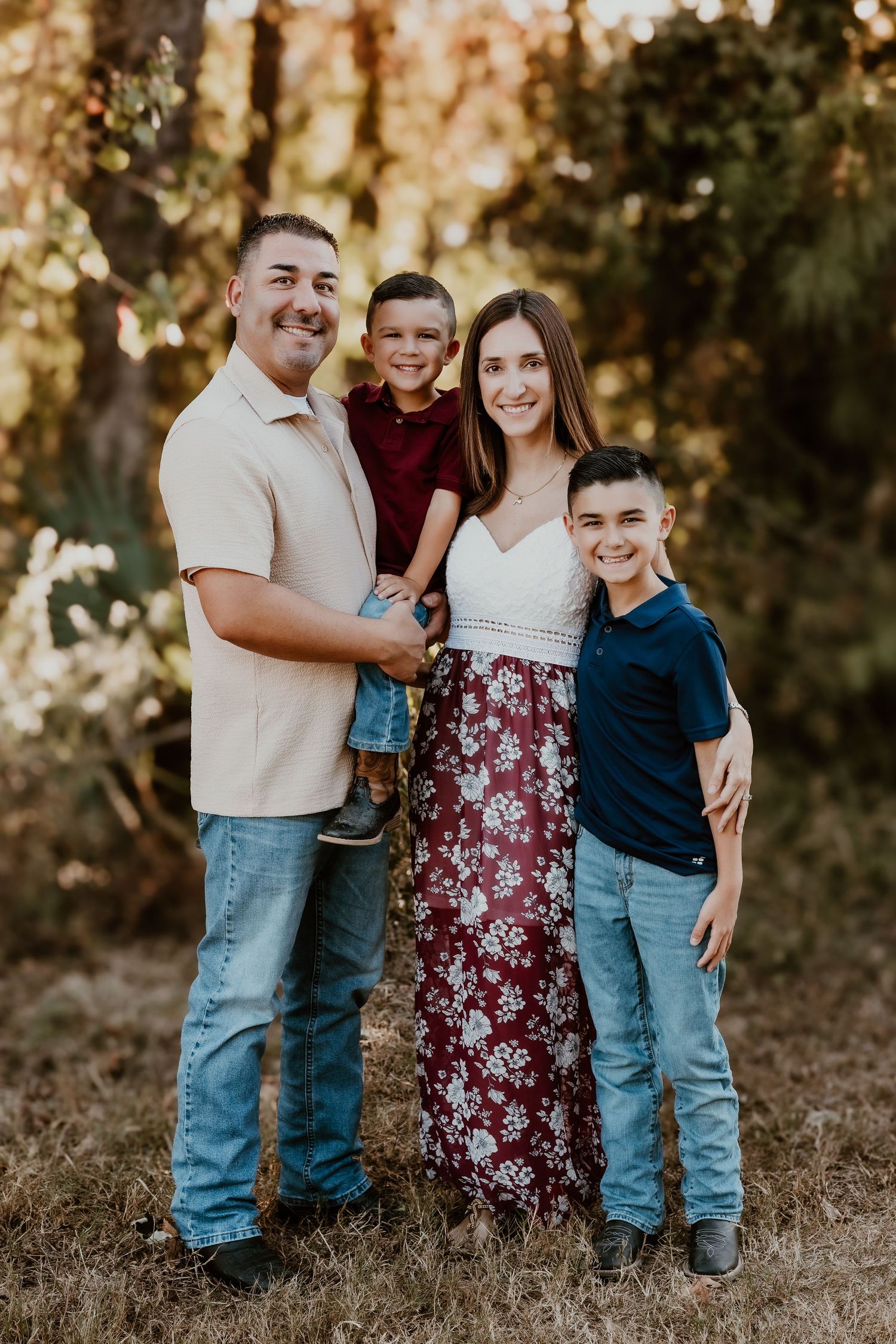 A family of four smiles at the camera while standing together in a natural outdoor setting at sunset.
