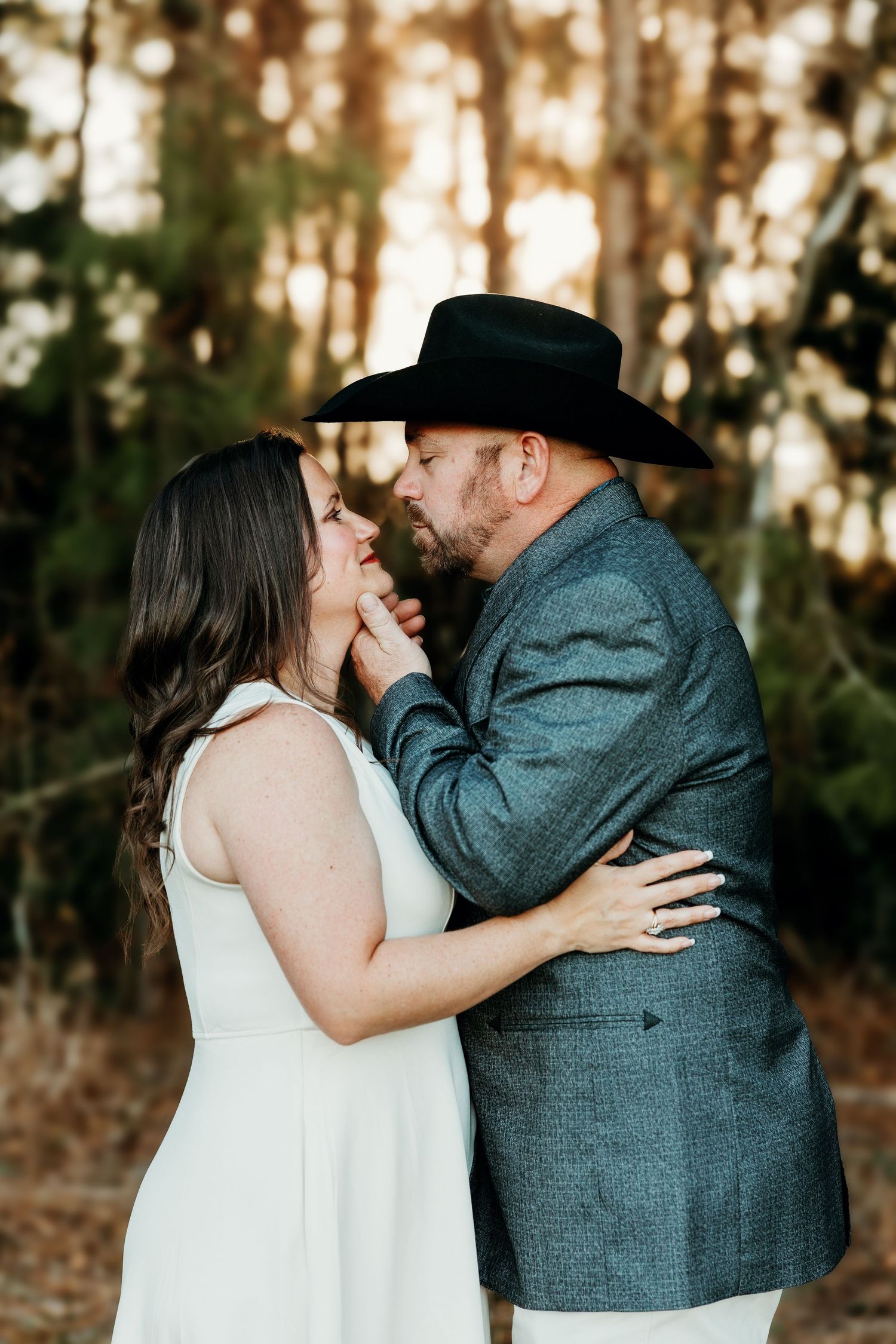 A couple shares romantic kisses in a wooded setting during sunset while wearing formal wedding attire.