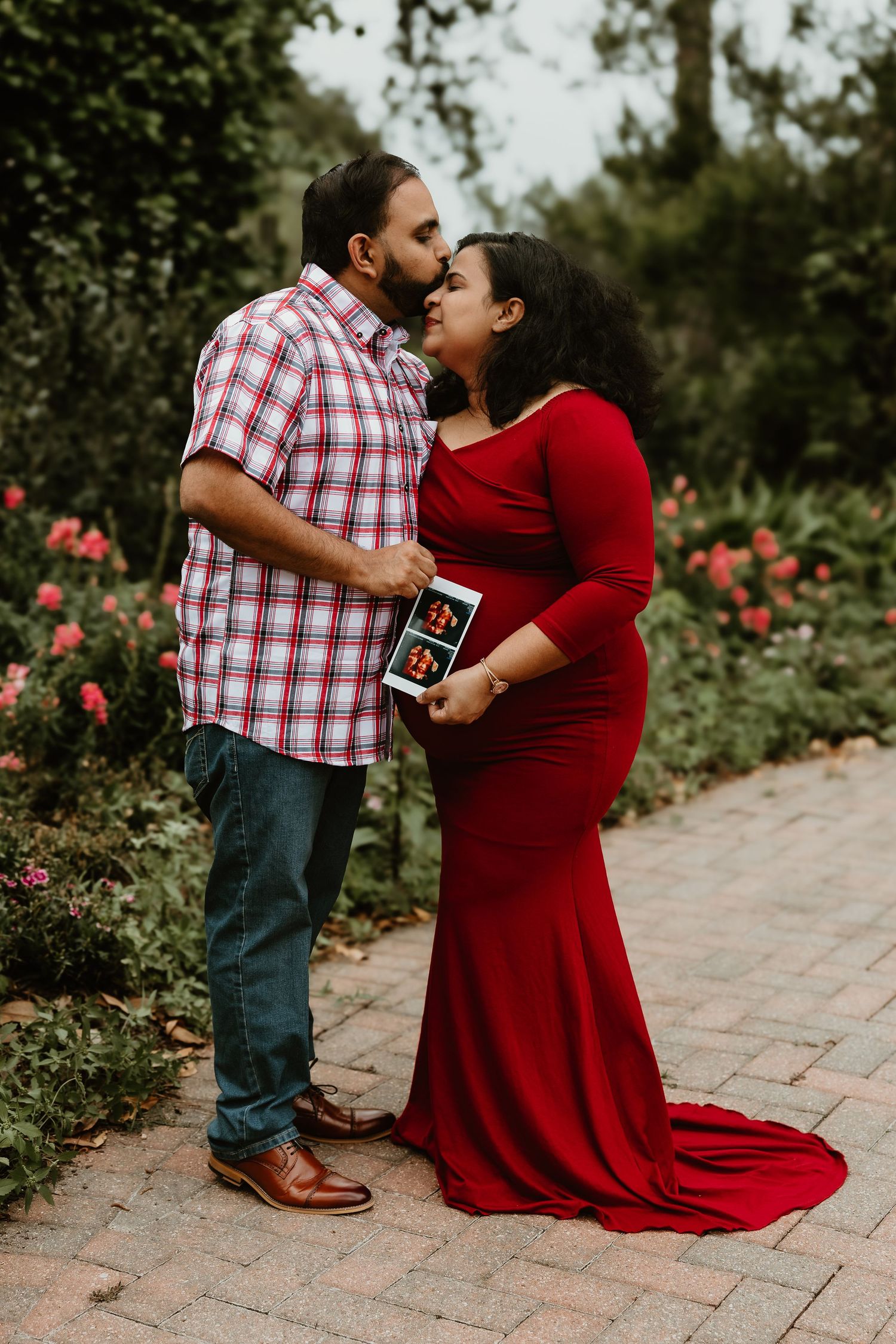 Pregnant couple sharing a kiss in a garden, woman in red maternity gown holding ultrasound photo, surrounded by roses.