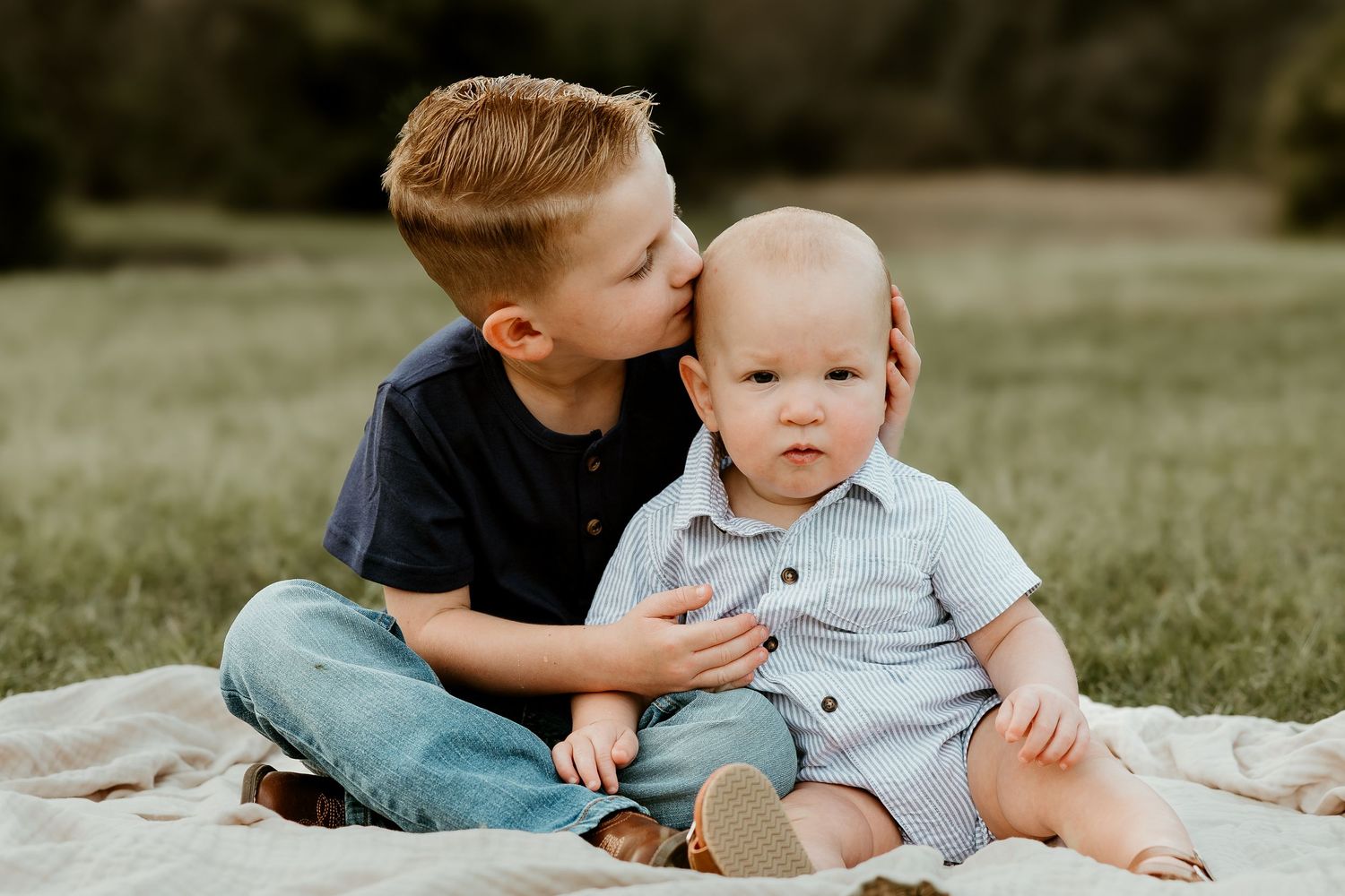 Two young children sit together on a blanket outdoors, with one giving the other a sweet kiss on the head.