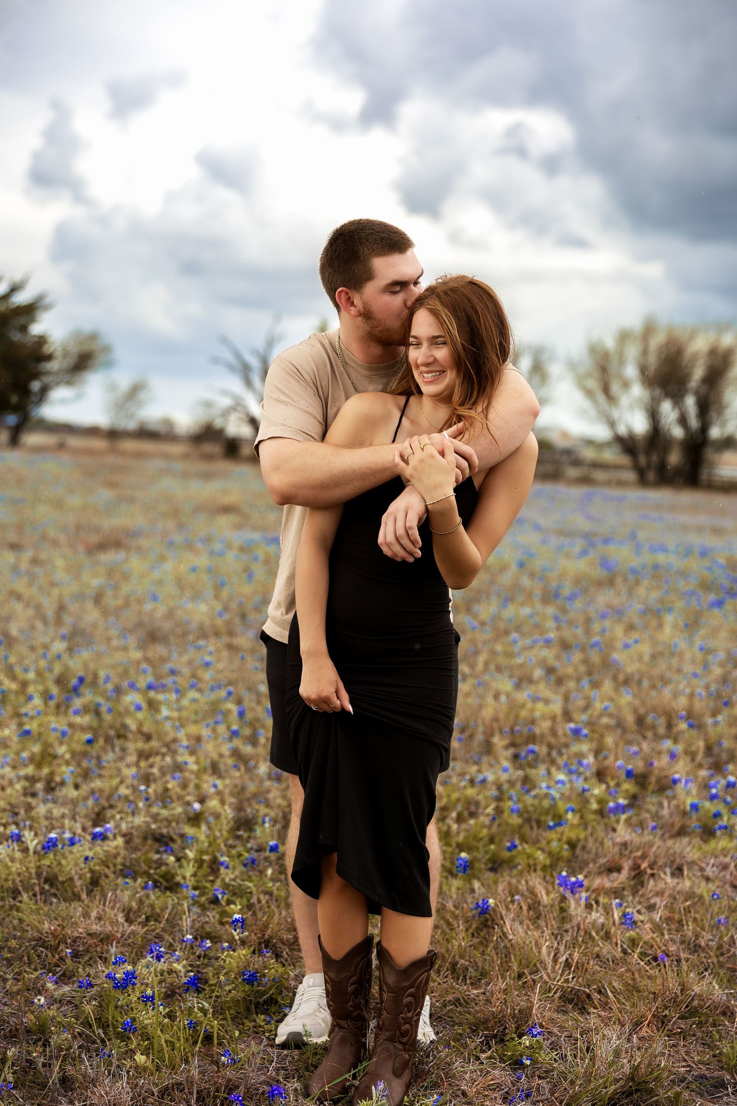 Couple embracing in a blue wildflower field, man kissing woman's head, she wears black dress and cowboy boots.