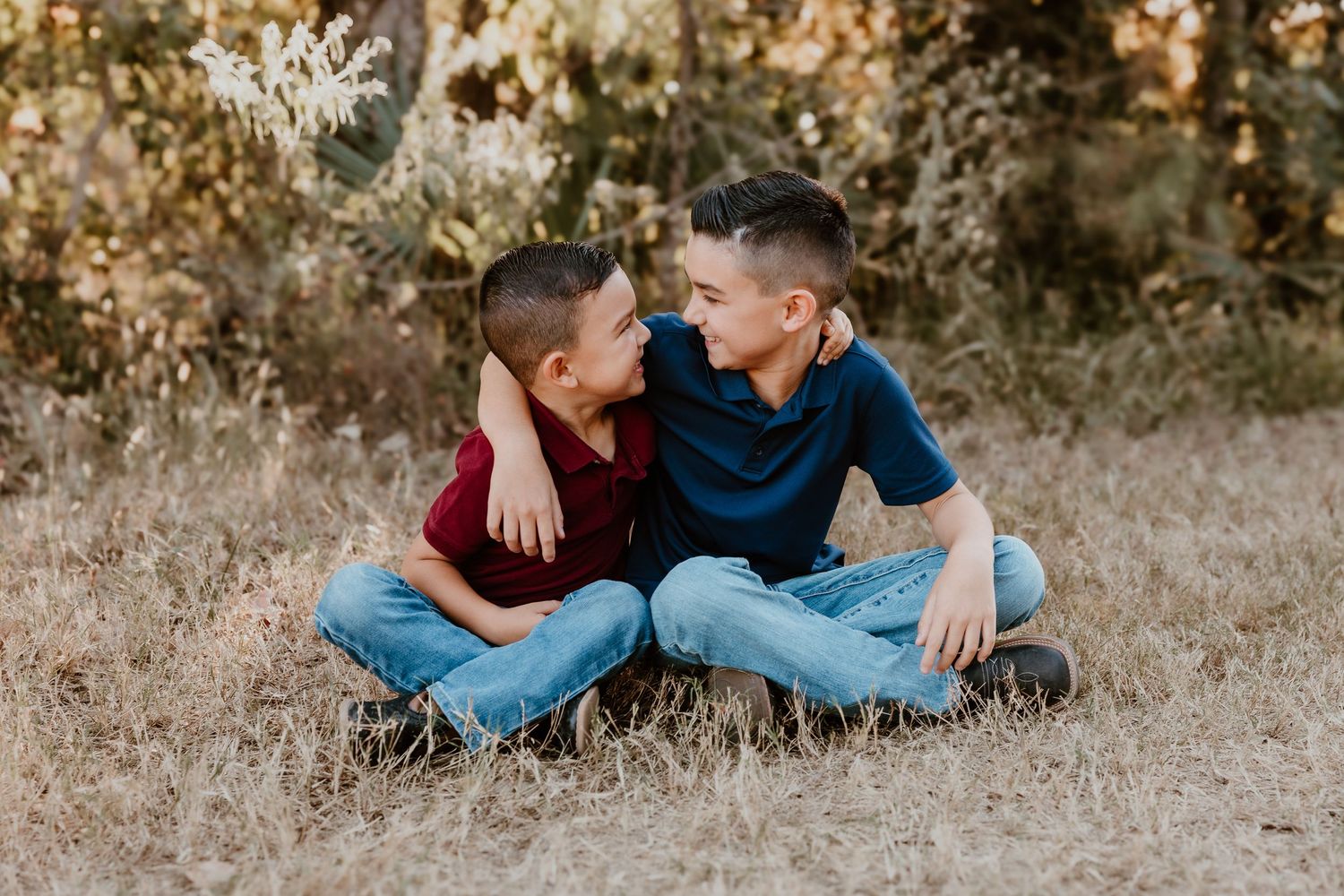 Two boys share a candid laughing moment while sitting cross-legged on dry grass.