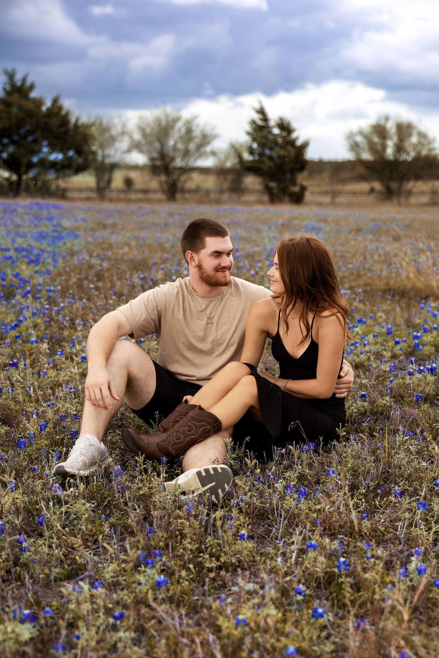 Couple sitting together in a blooming blue wildflower field, smiling at each other on a cloudy day.