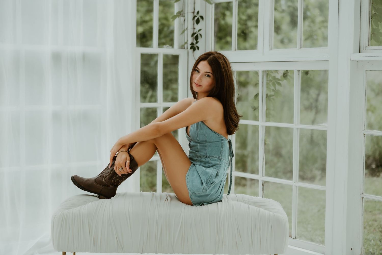 A model in a blue denim dress poses on a white surface near bright windows.