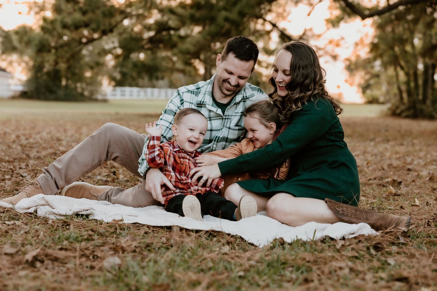 A family sits together on a blanket outdoors during autumn, laughing and embracing on fallen leaves at sunset.