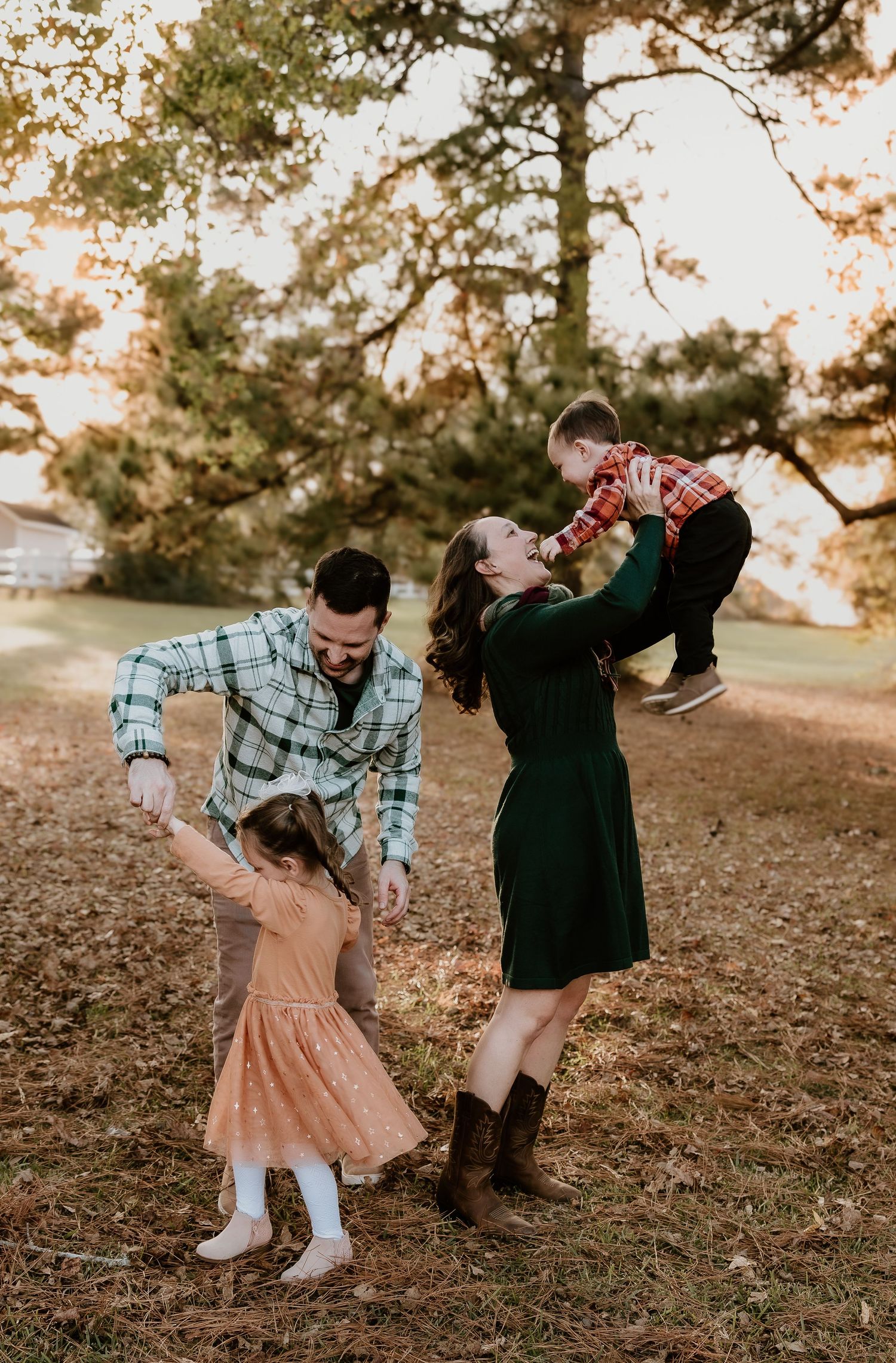 A family plays together outdoors during golden hour in autumn, with pine trees and fallen leaves in the background.