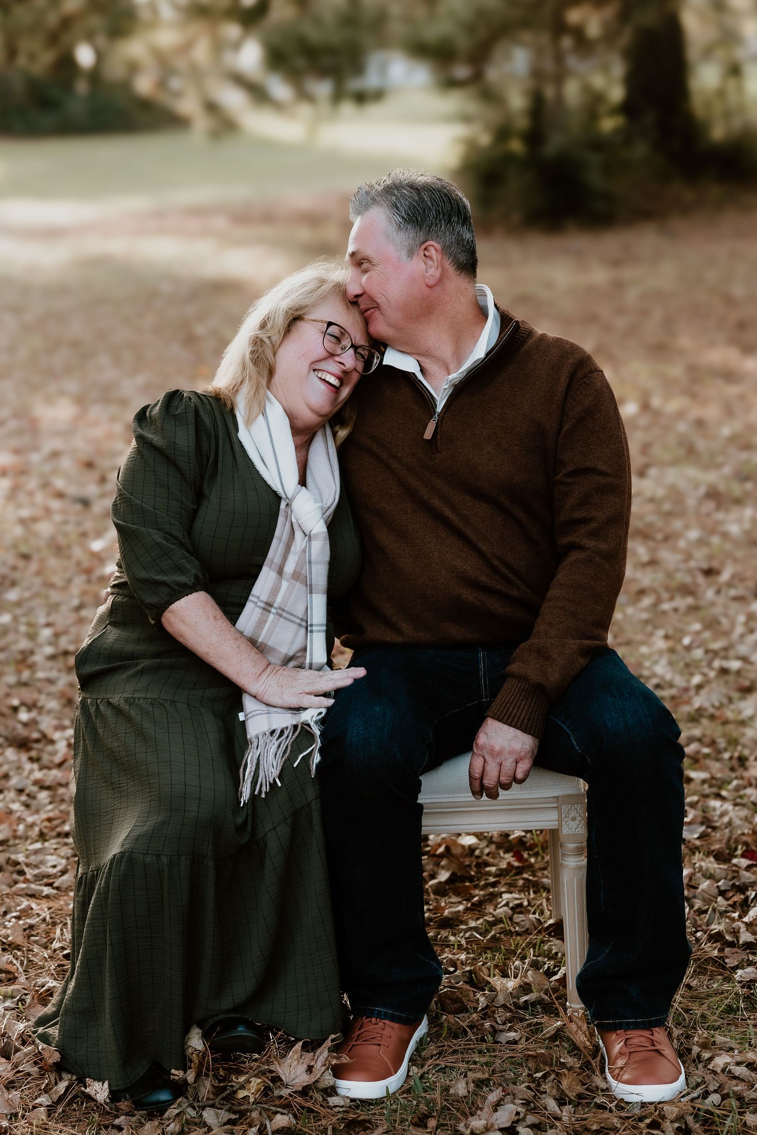 A couple shares a tender moment while sitting on a chair outdoors during autumn, surrounded by fallen leaves.