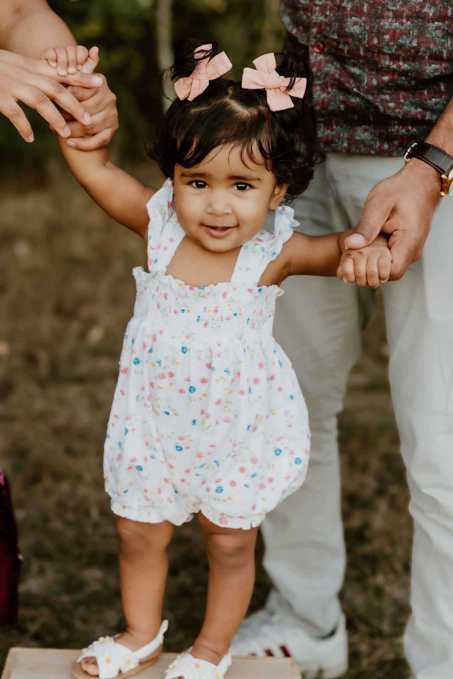 A small child in a white floral romper holds hands with someone while standing outdoors.