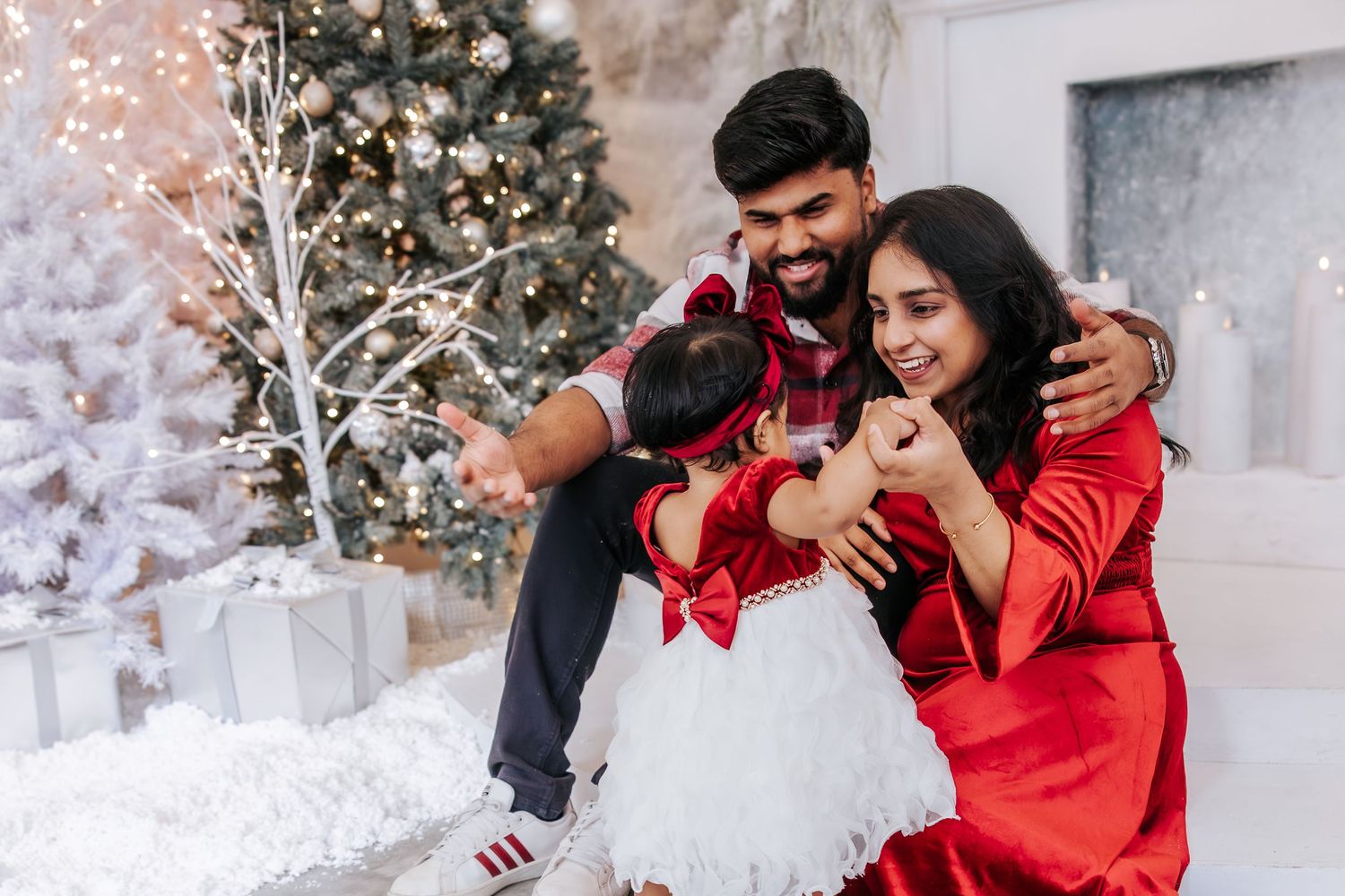 A romantic winter photo shoot with a couple in red outfits surrounded by snowy Christmas trees and white decor.