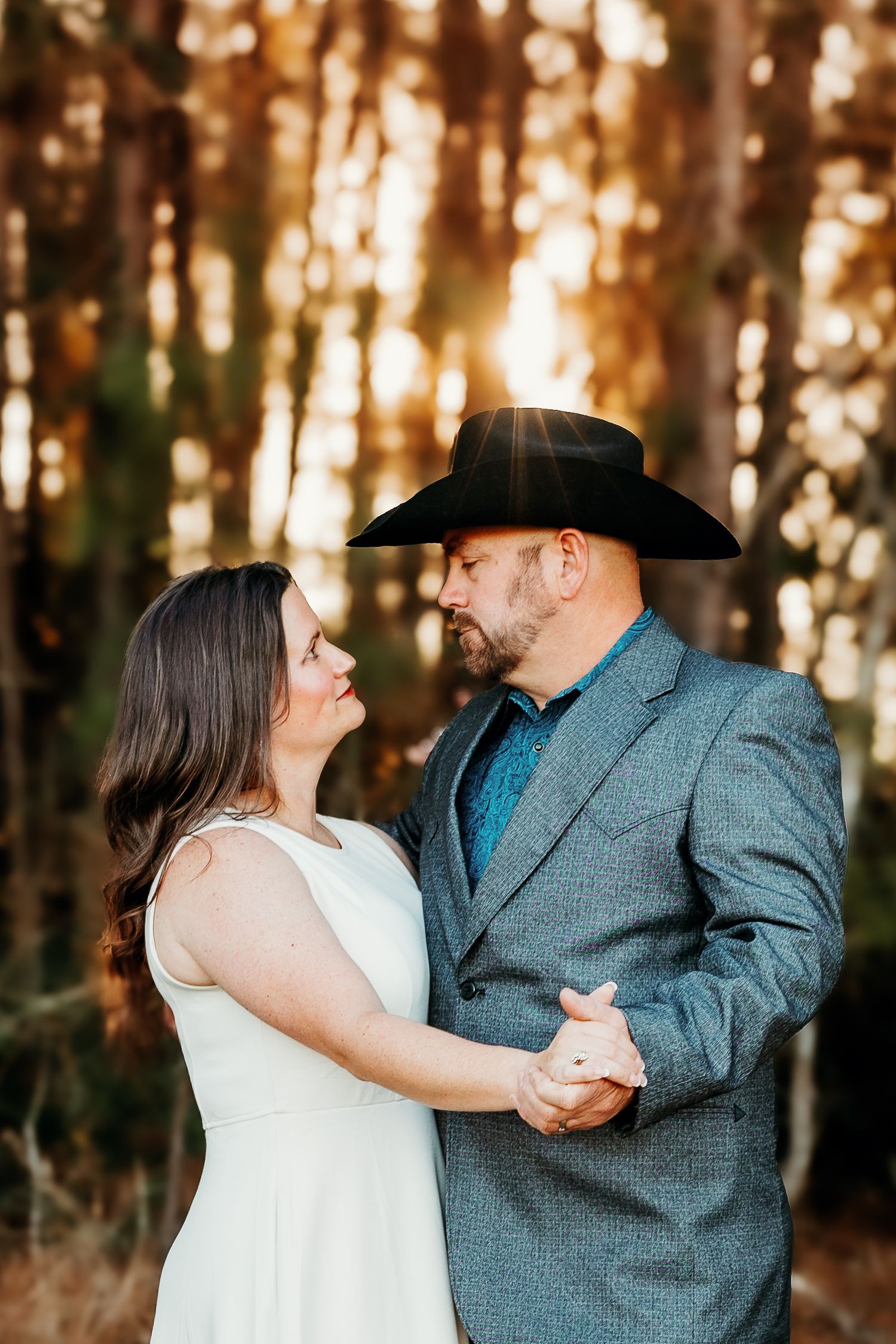A couple dances together in a forest at sunset, with the woman in white dress and man wearing cowboy hat and blazer.