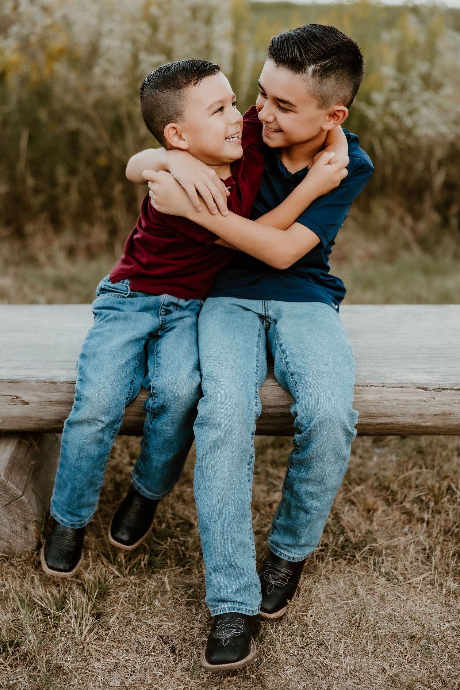 Two siblings share a playful moment sitting on a wooden fence in casual clothing.
