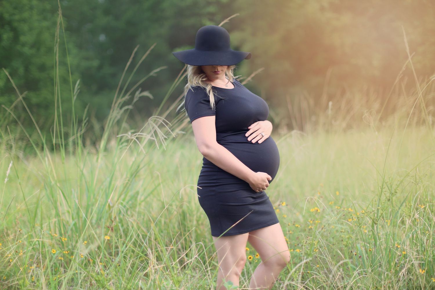 Person in black clothing and baseball cap standing in tall grass during golden hour.