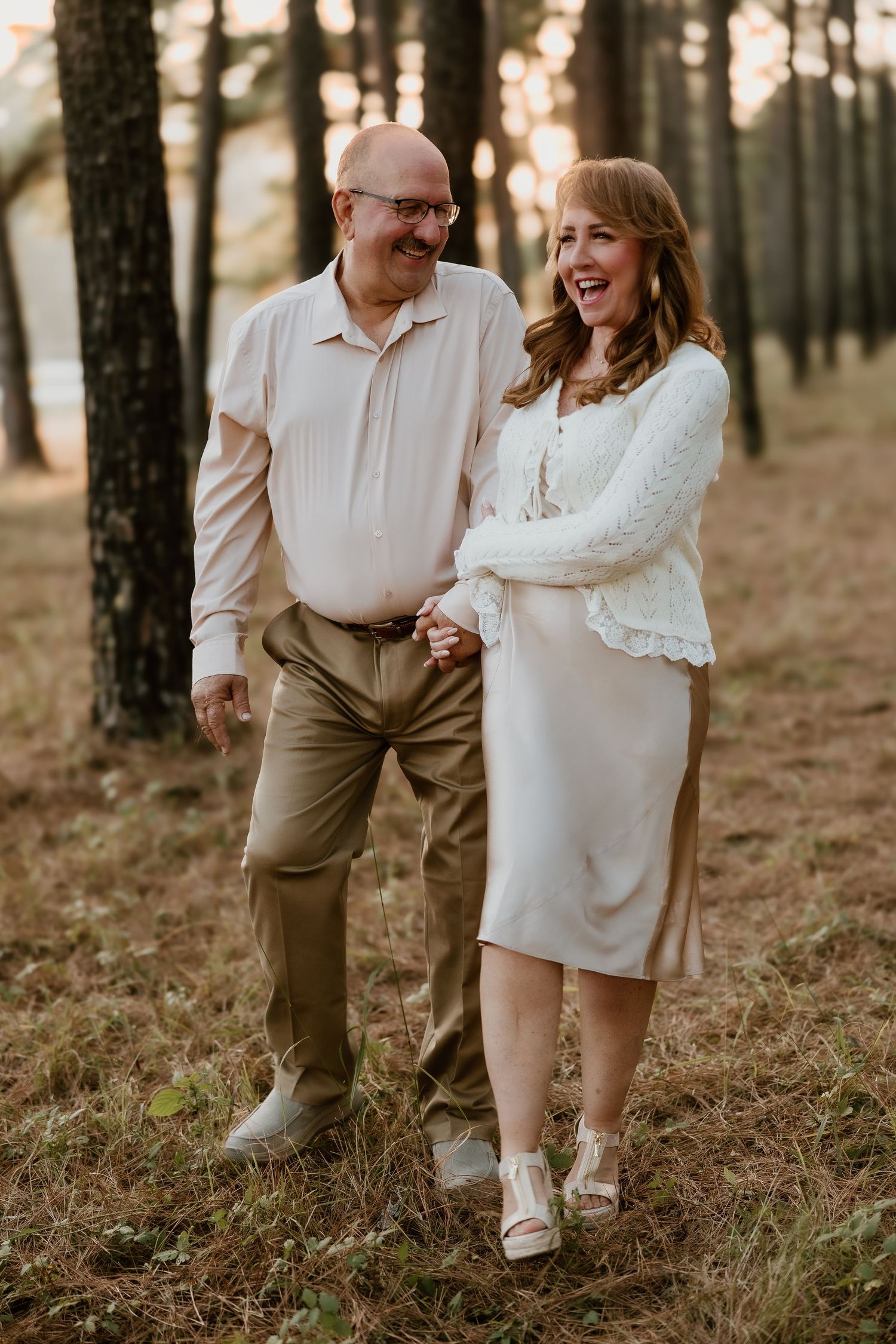 A couple walks hand in hand through a sunlit forest wearing light colored outfits and sharing joyful laughter.