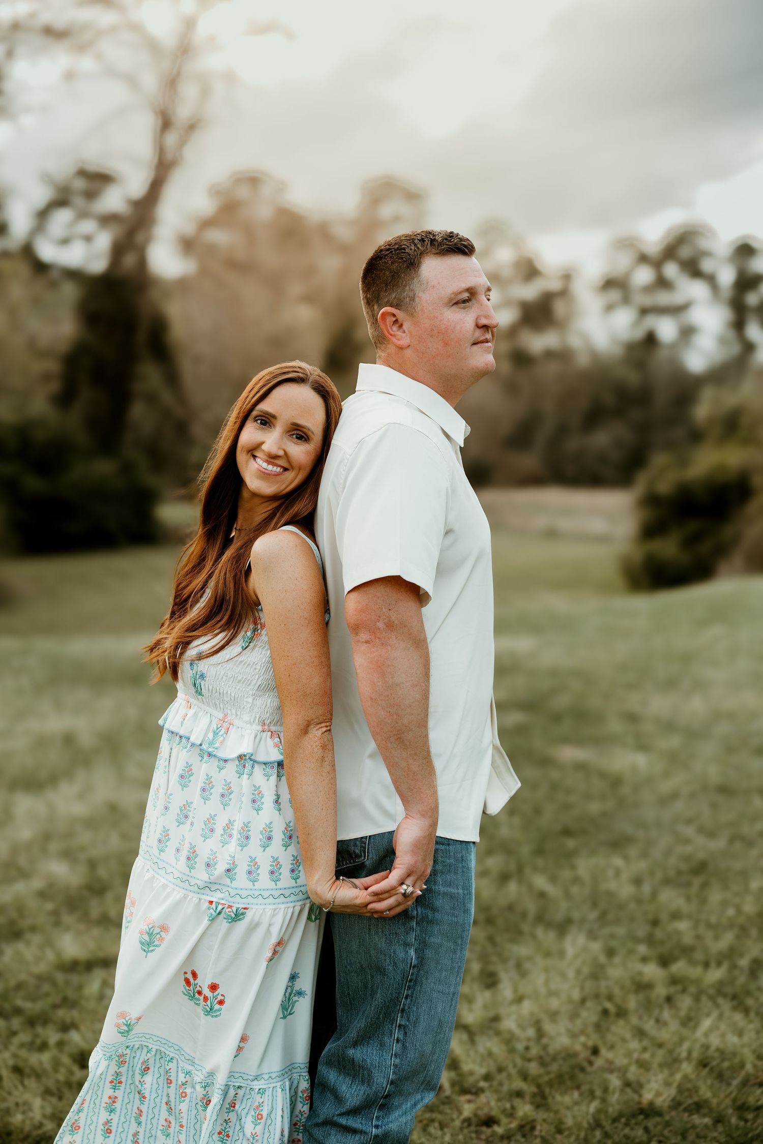 A romantic couple stands together in a grassy field during golden hour wearing white and blue casual attire.