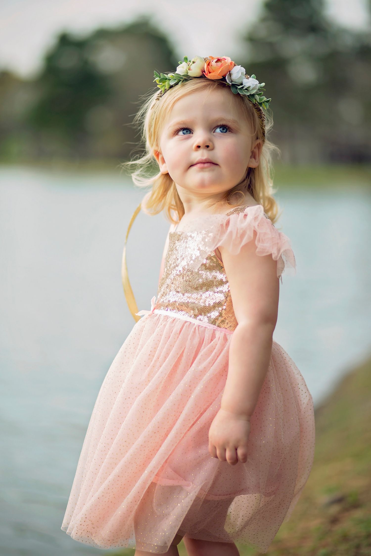 A toddler in a peach tulle dress and floral crown stands near water during a photo shoot.