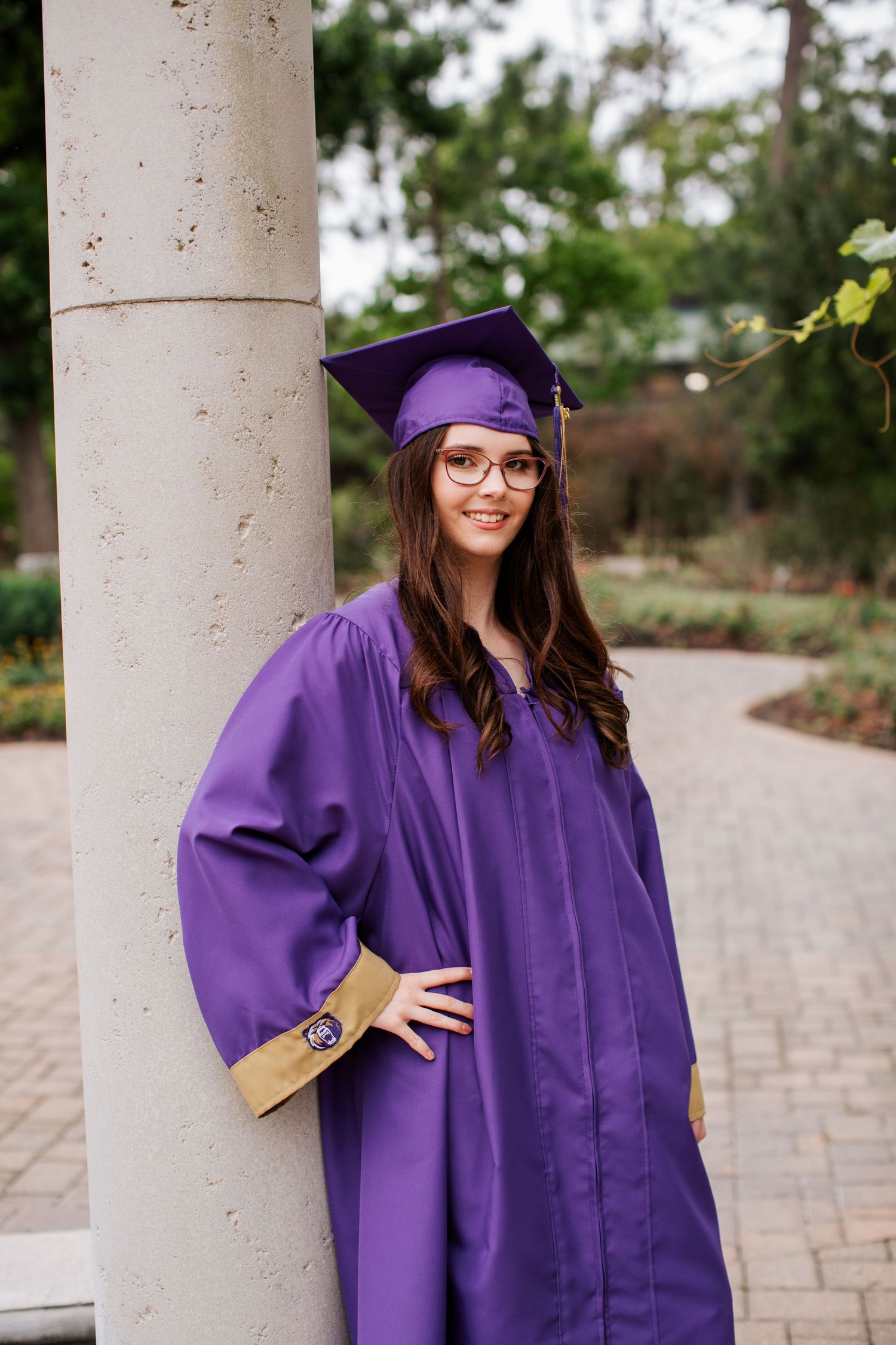 Graduate in purple cap and gown posing by stone column on brick pathway.