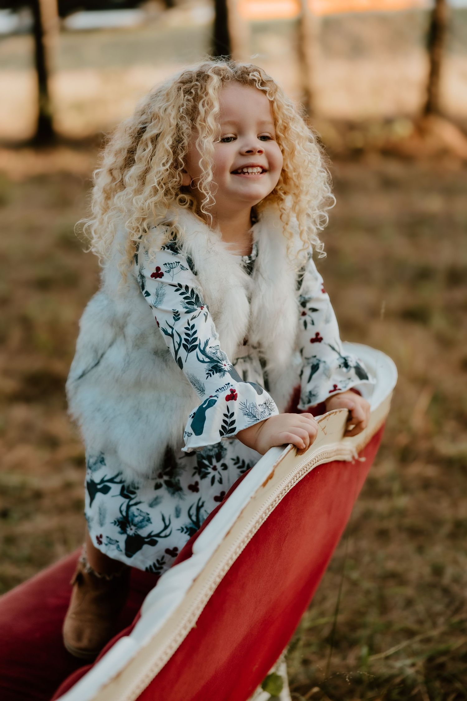 Child with curly blonde hair wearing a floral dress plays joyfully on a red outdoor slide in autumn light.