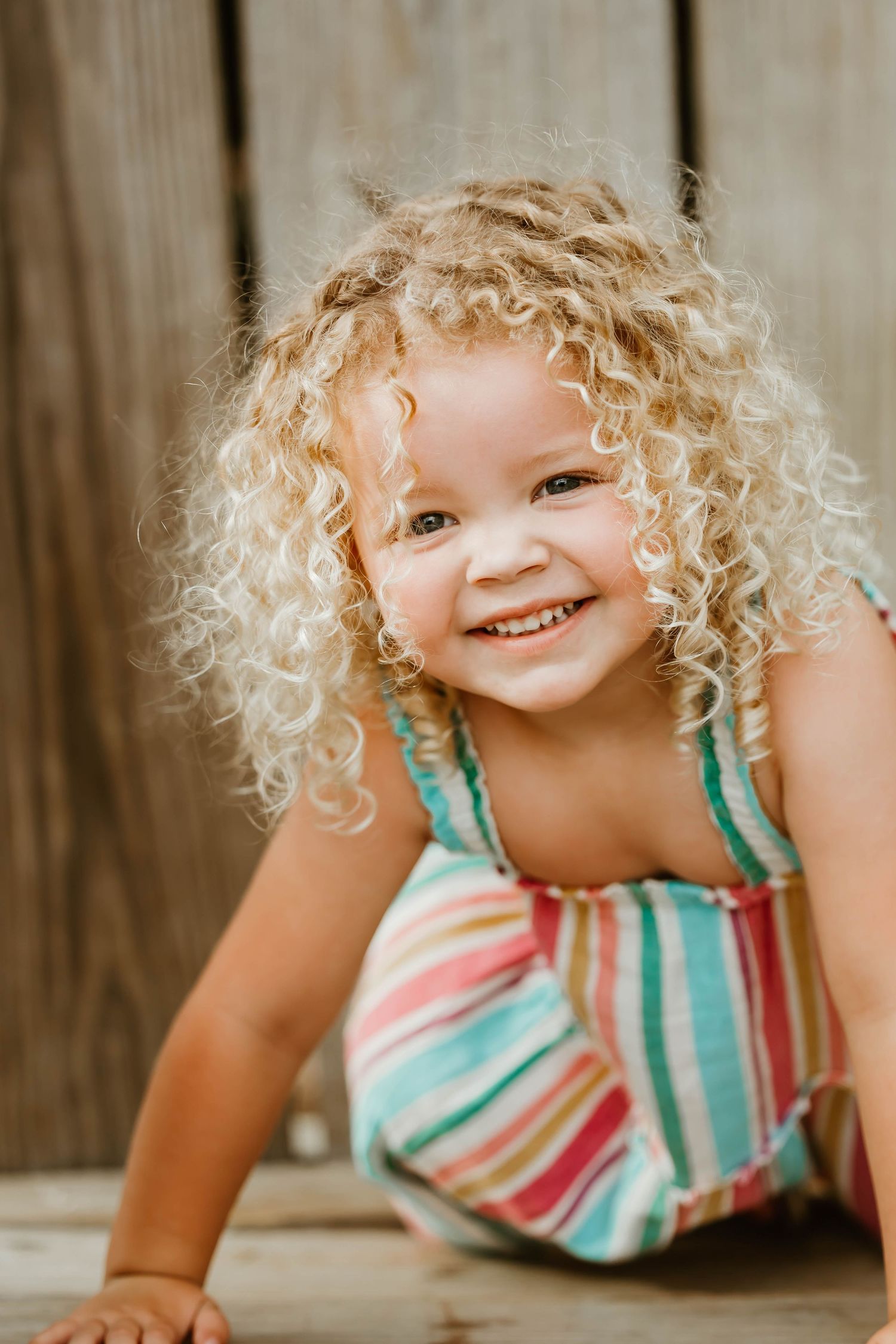 A child with curly blonde hair smiles playfully in a striped summer dress.
