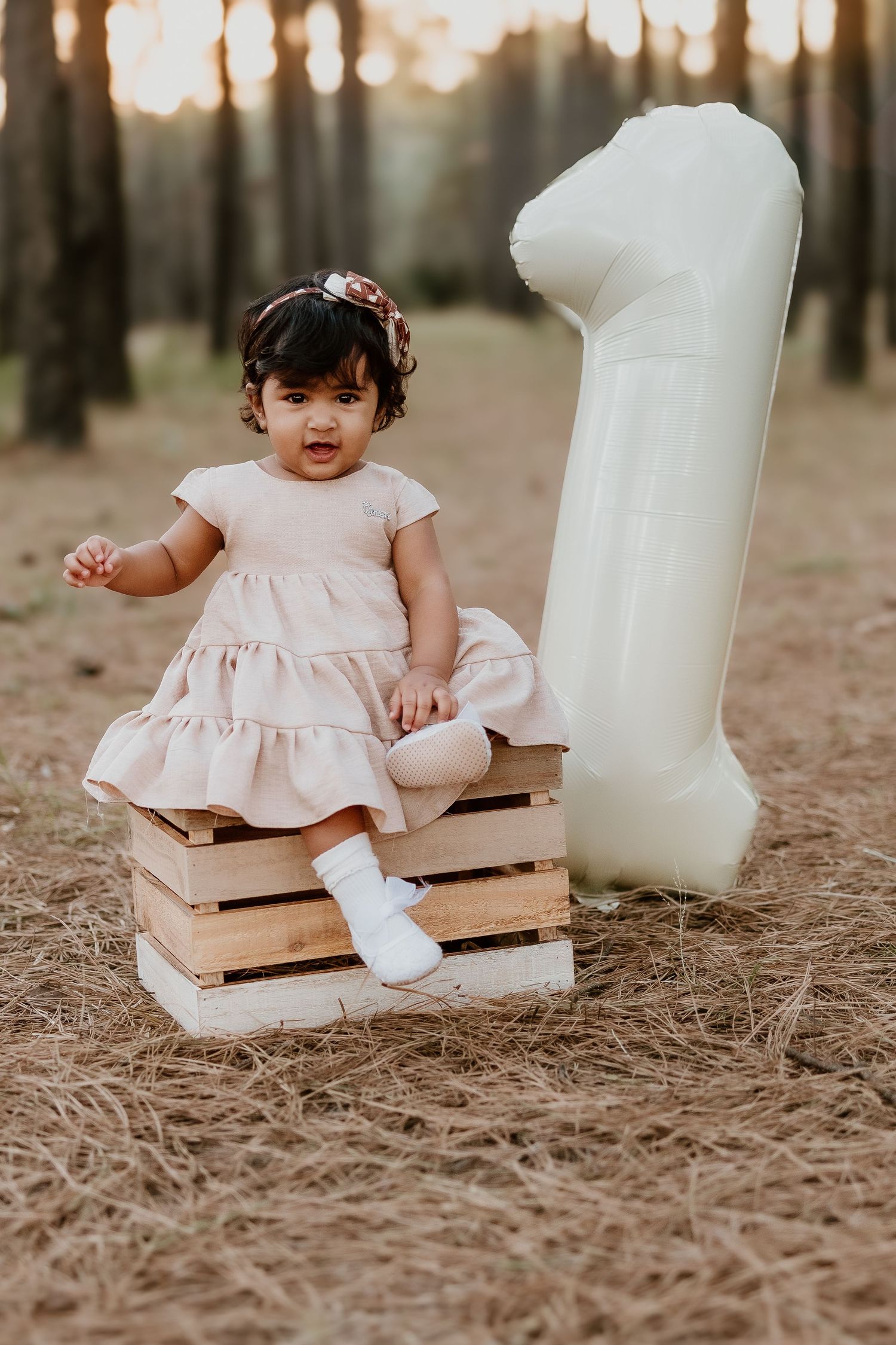 Baby in pink dress sits on wooden crate in pine forest with dreamy sunset lighting in background.