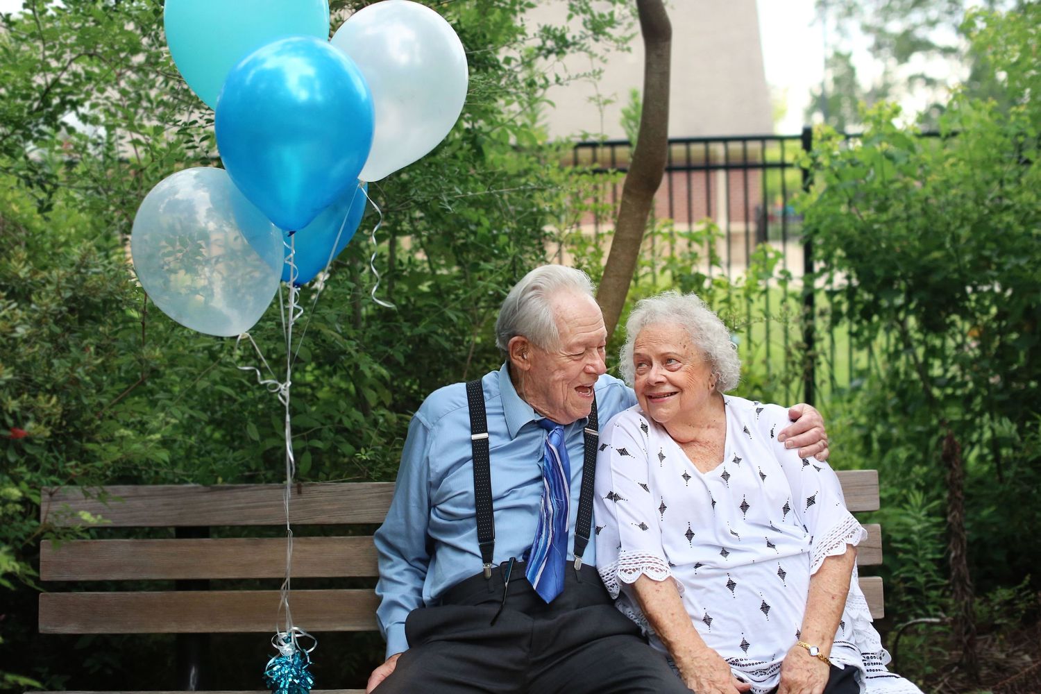 Elderly couple sits on bench with blue and white balloons during anniversary photo session.