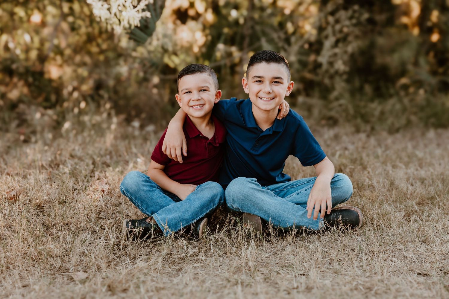 Two boys in casual wear sit together smiling on dry grass during sunset.