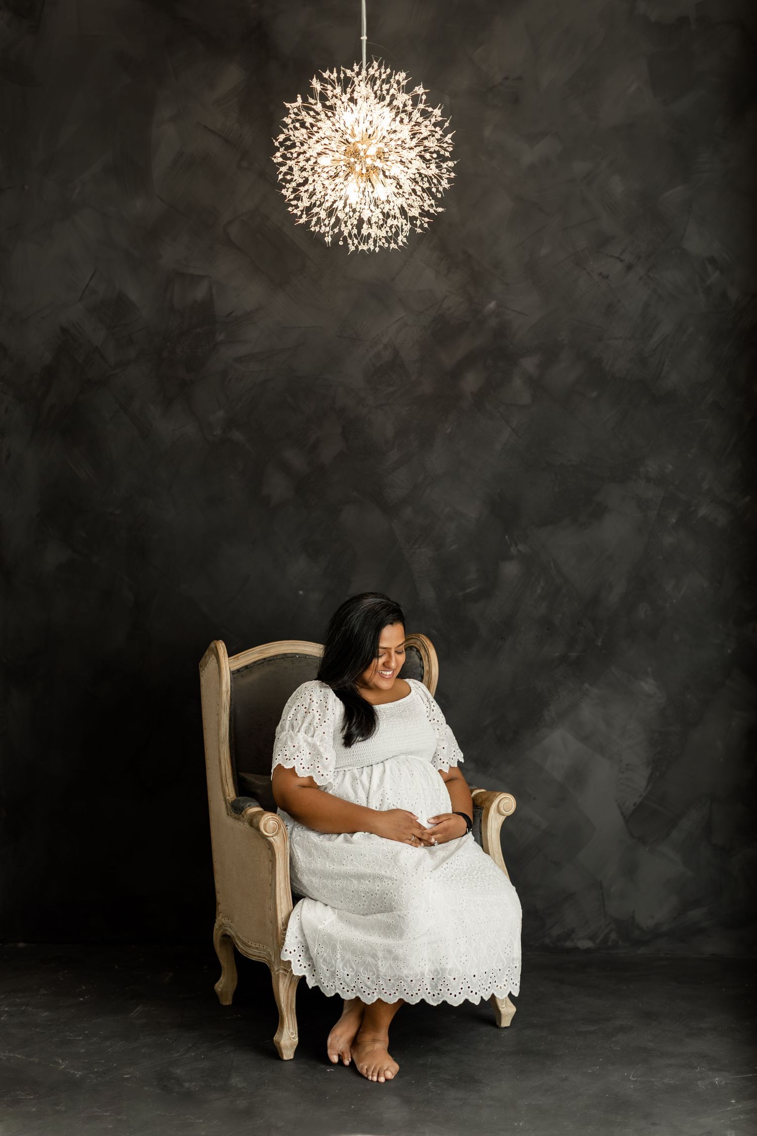 Pregnant woman in white lace dress sitting in beige armchair under chandelier against dark gray studio backdrop.