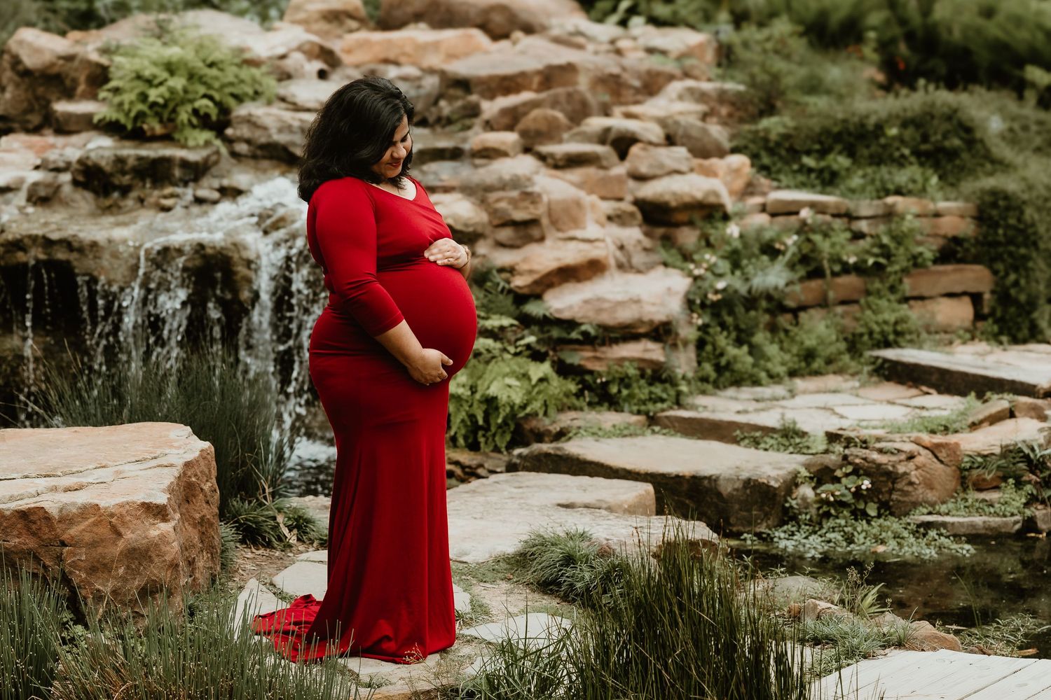 Pregnant woman in red gown poses by waterfall and rocks in lush garden during maternity photo session.