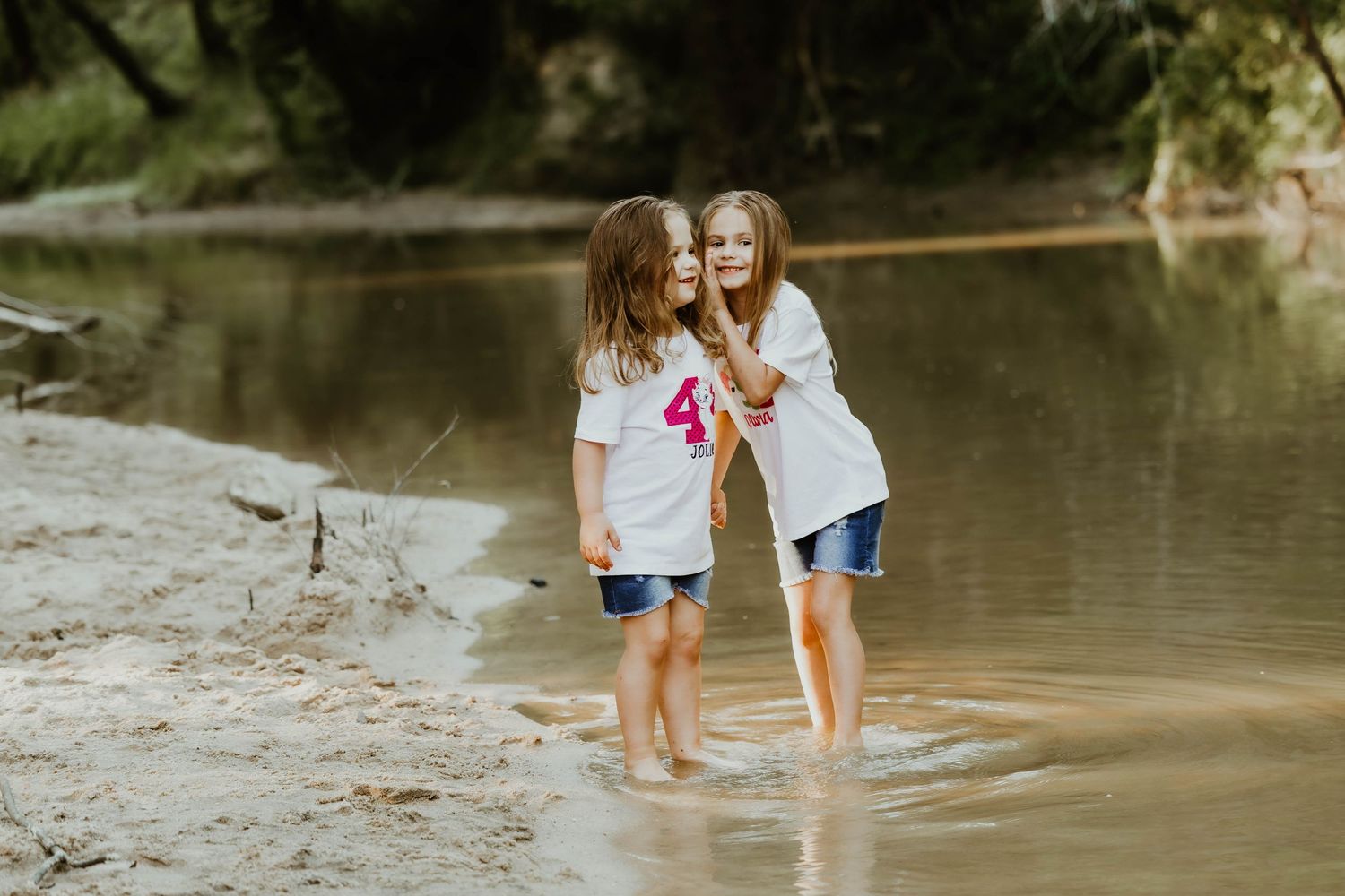 Two children in matching white shirts explore the muddy shoreline of a creek.