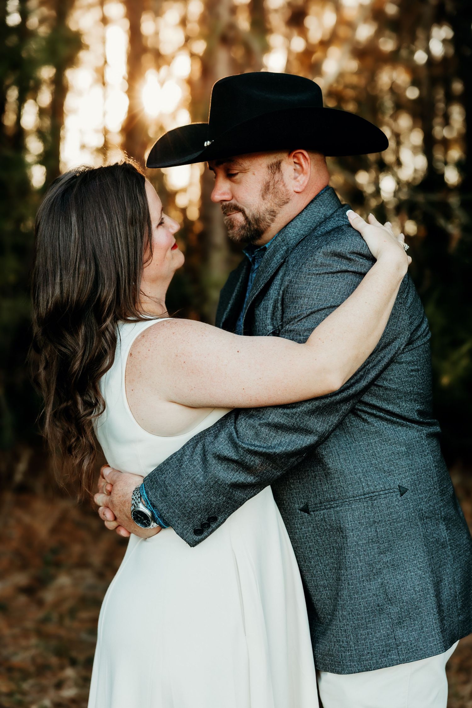 A romantic couple embraces in a sunlit forest setting, with the bride in white and groom in a gray suit and cowboy hat.