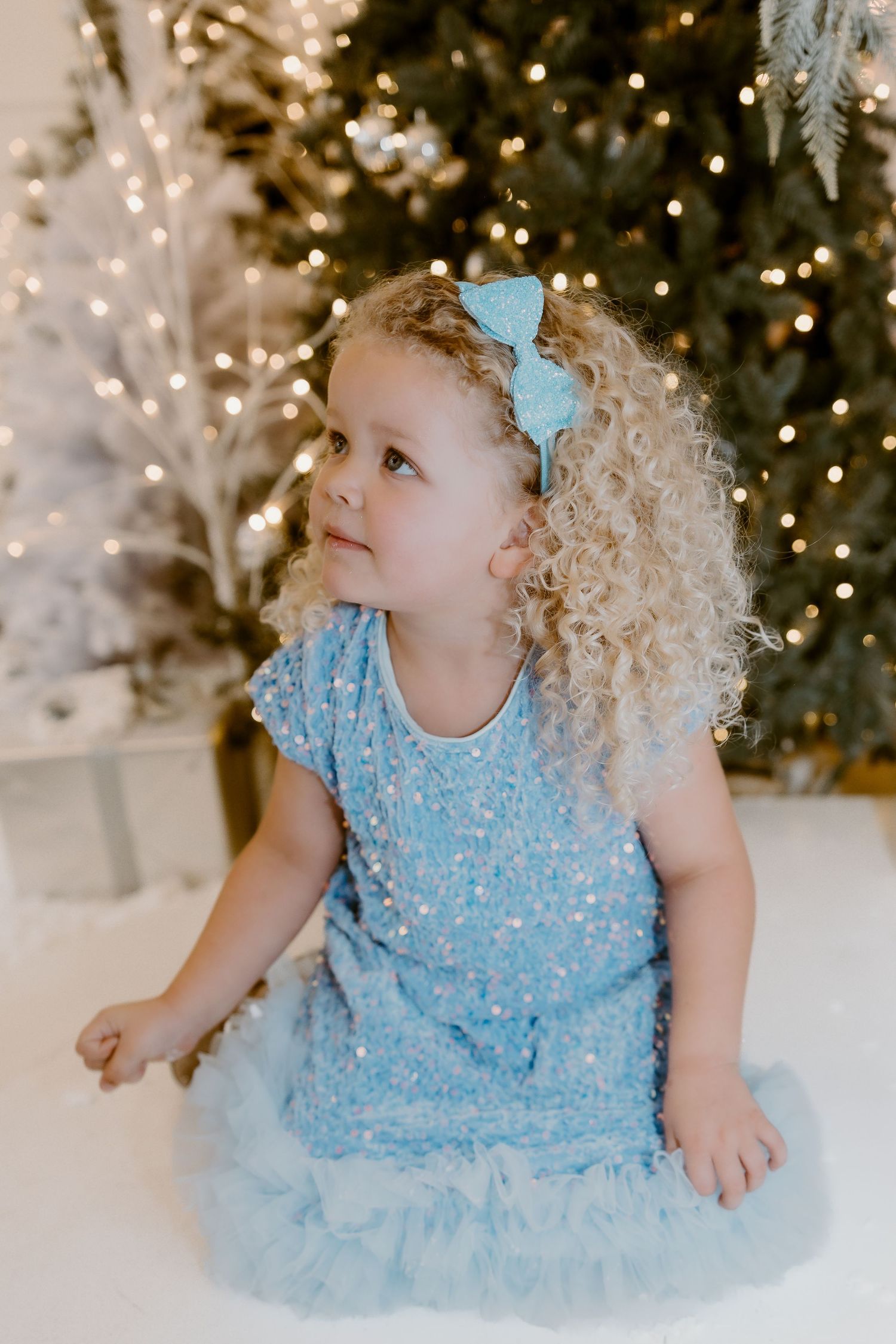 A child in a light blue dress sits on a white floor with twinkling Christmas lights in the background.