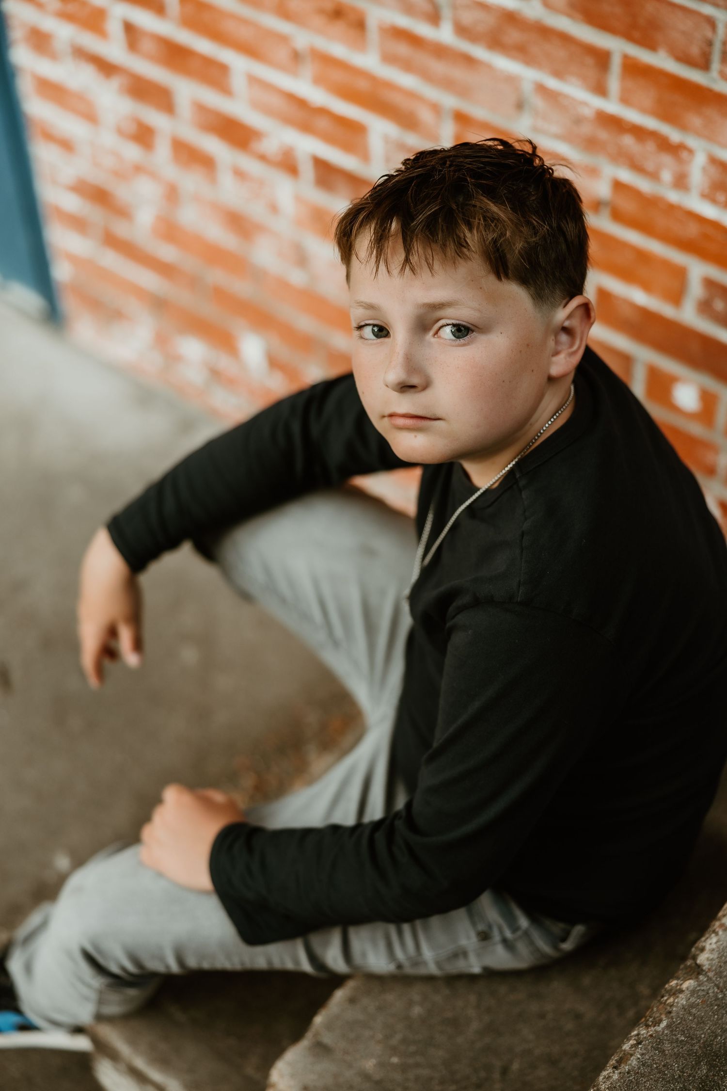 A casual portrait against a brick wall shows a youth in a black long sleeve shirt and gray pants.