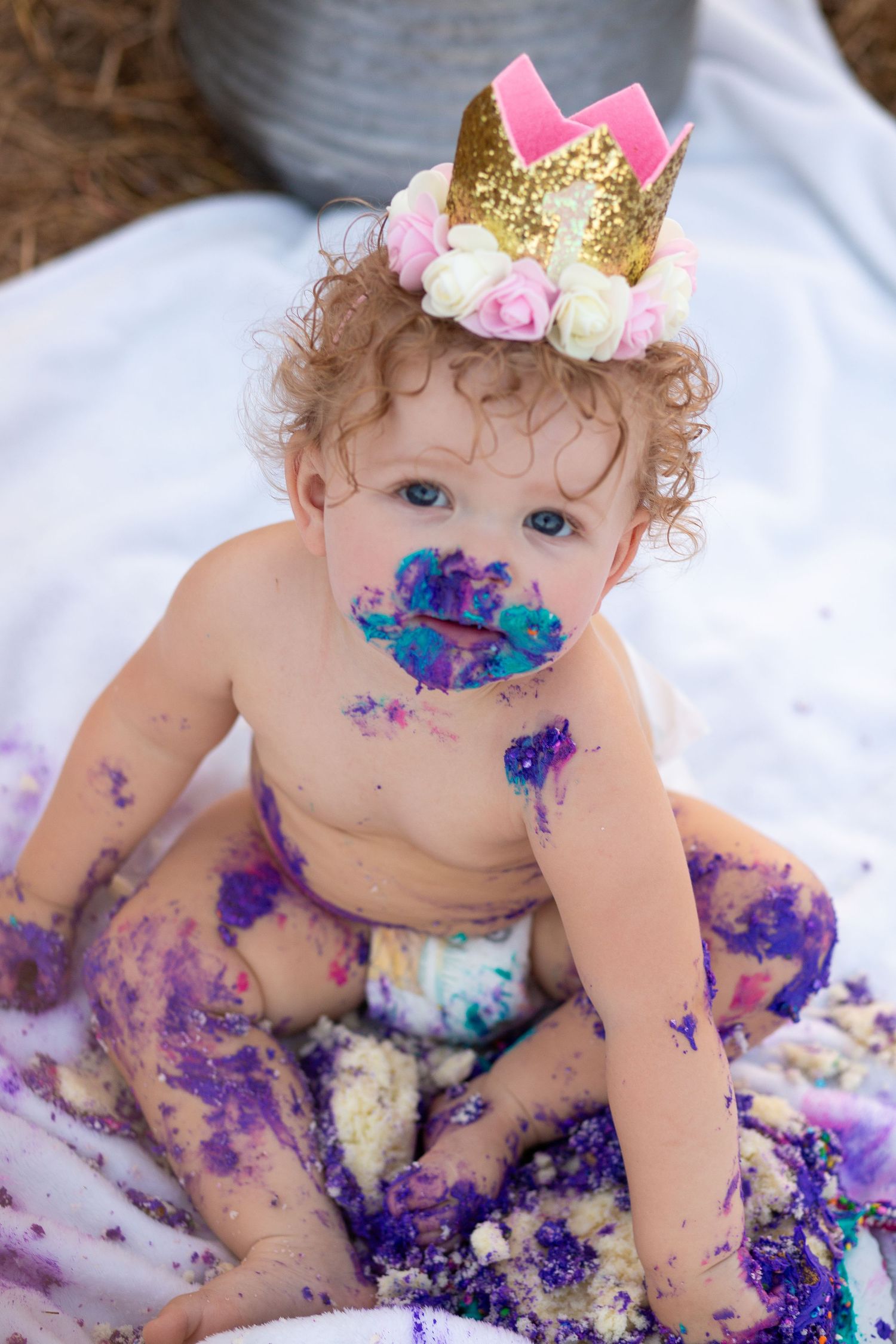 Baby wearing gold crown gets messy with purple cake during smash cake photo session.