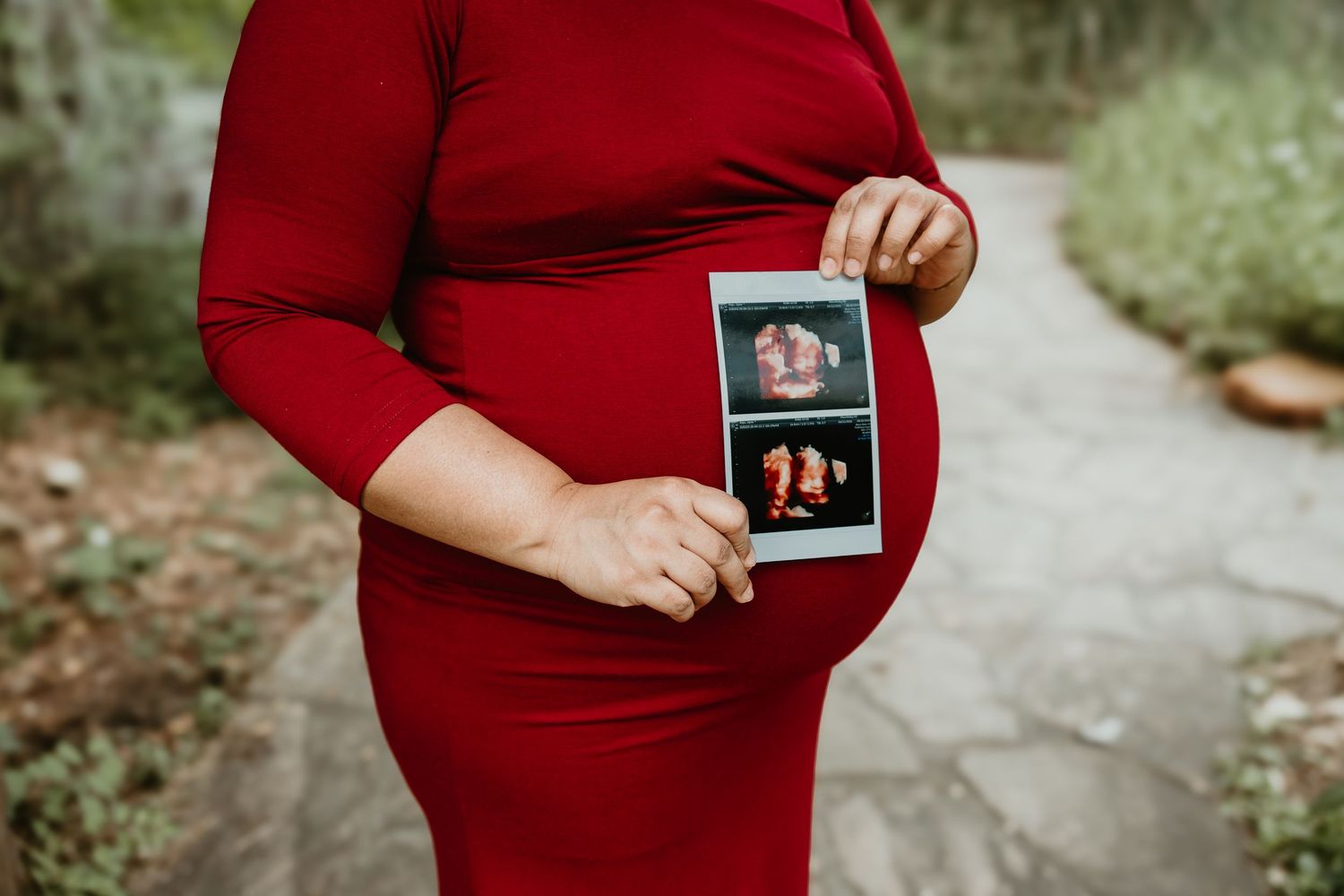 Pregnant woman in red dress holding ultrasound photos outdoors on a stone path.