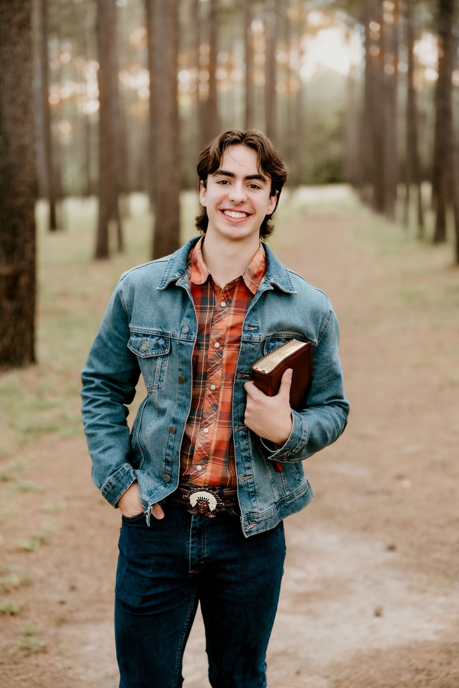 Smiling young man in denim jacket holding a Bible on a forest path lined with tall pine trees.