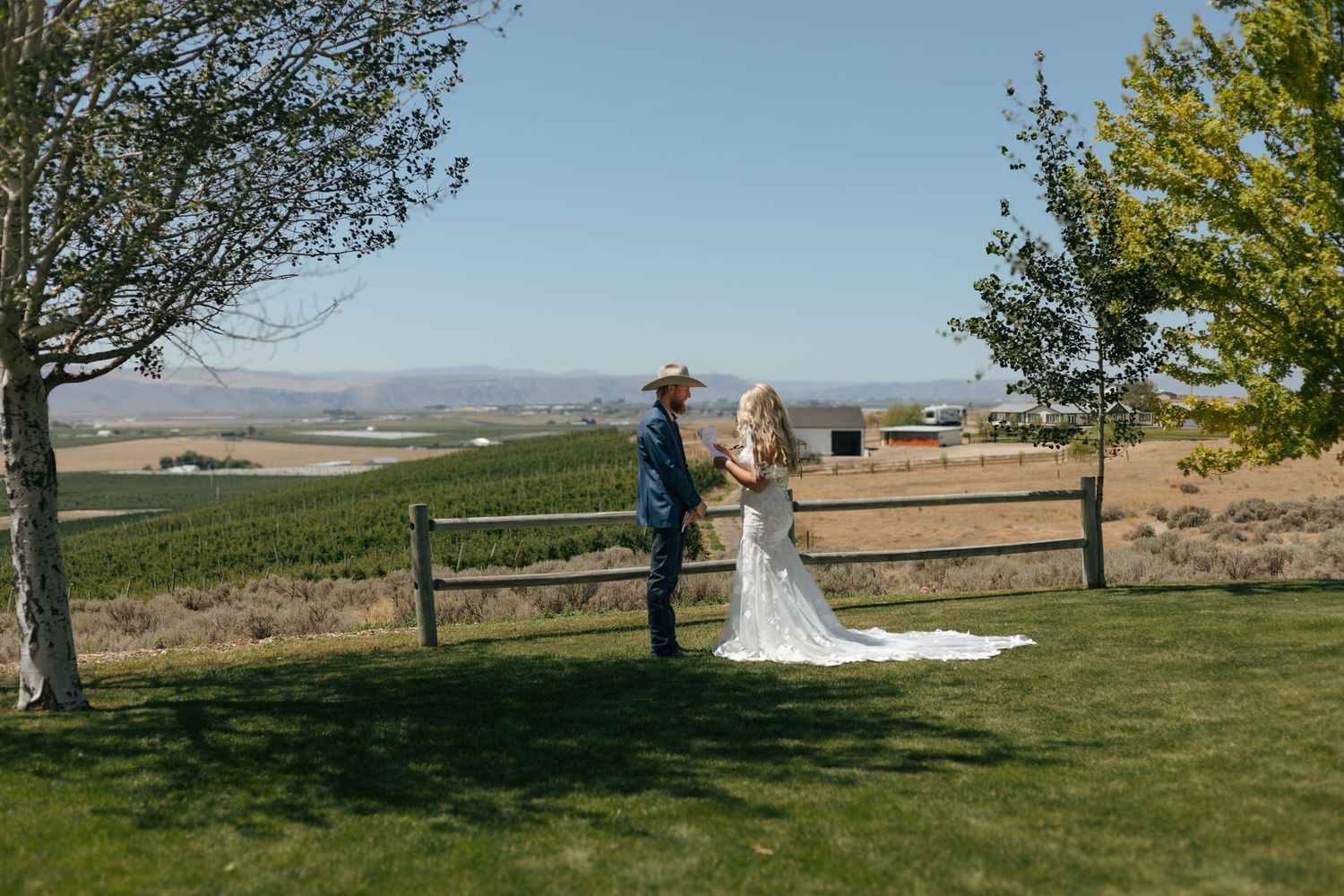 Bride and groom stand on green lawn at rustic vineyard wedding venue with scenic valley views.