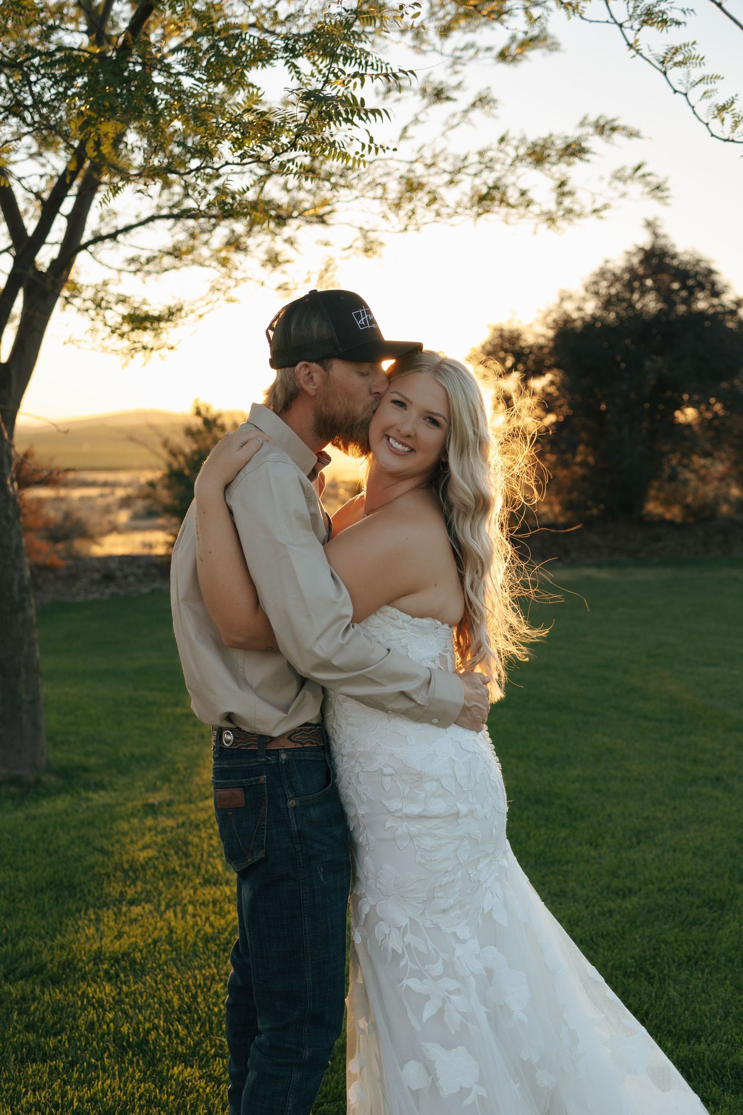 Bride in white lace gown embraced by groom in tan shirt and cap at golden hour outdoor wedding.