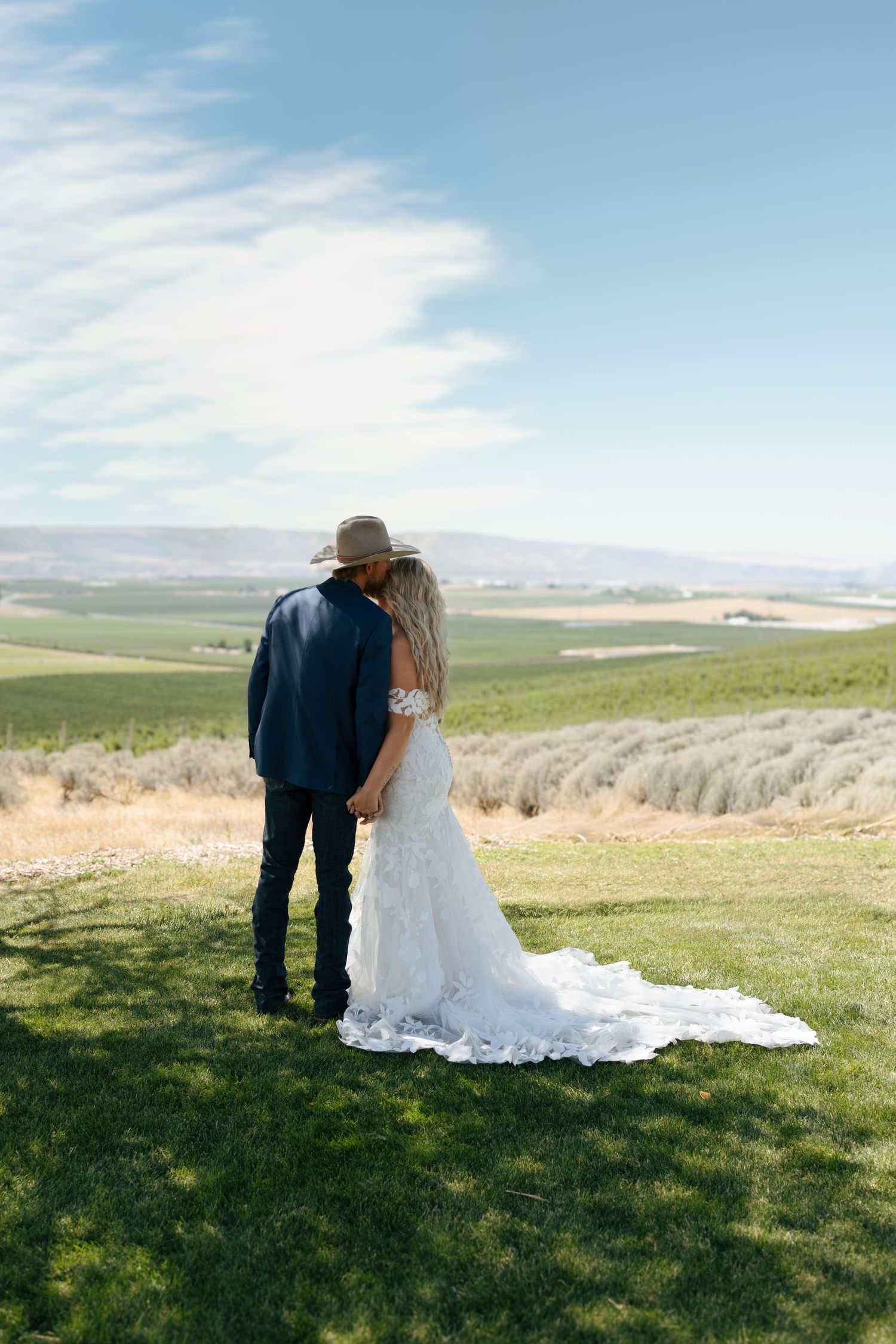Bride in lace gown and groom in cowboy hat hold hands overlooking scenic vineyard landscape on wedding day.