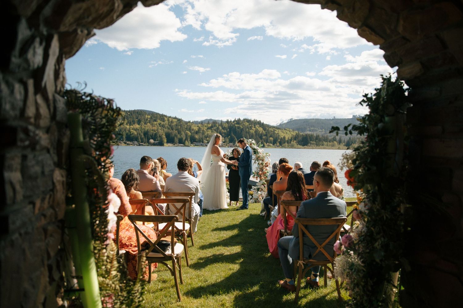 Outdoor lakeside wedding ceremony viewed through stone arch, guests seated on wooden chairs, couple at altar.