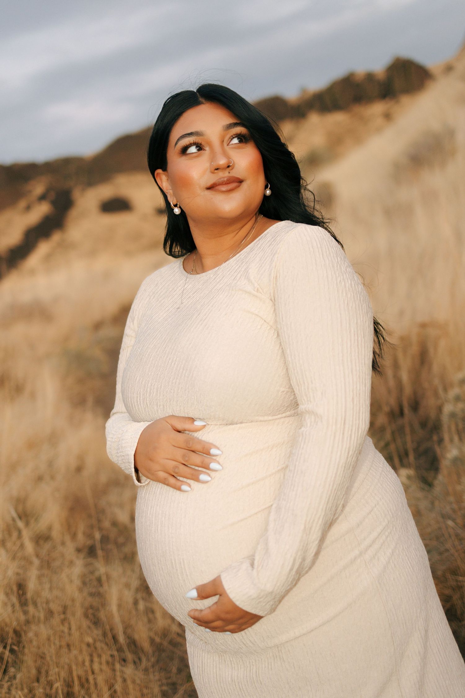 Pregnant woman in cream ribbed dress cradling baby bump outdoors in golden dry grass field at sunset.