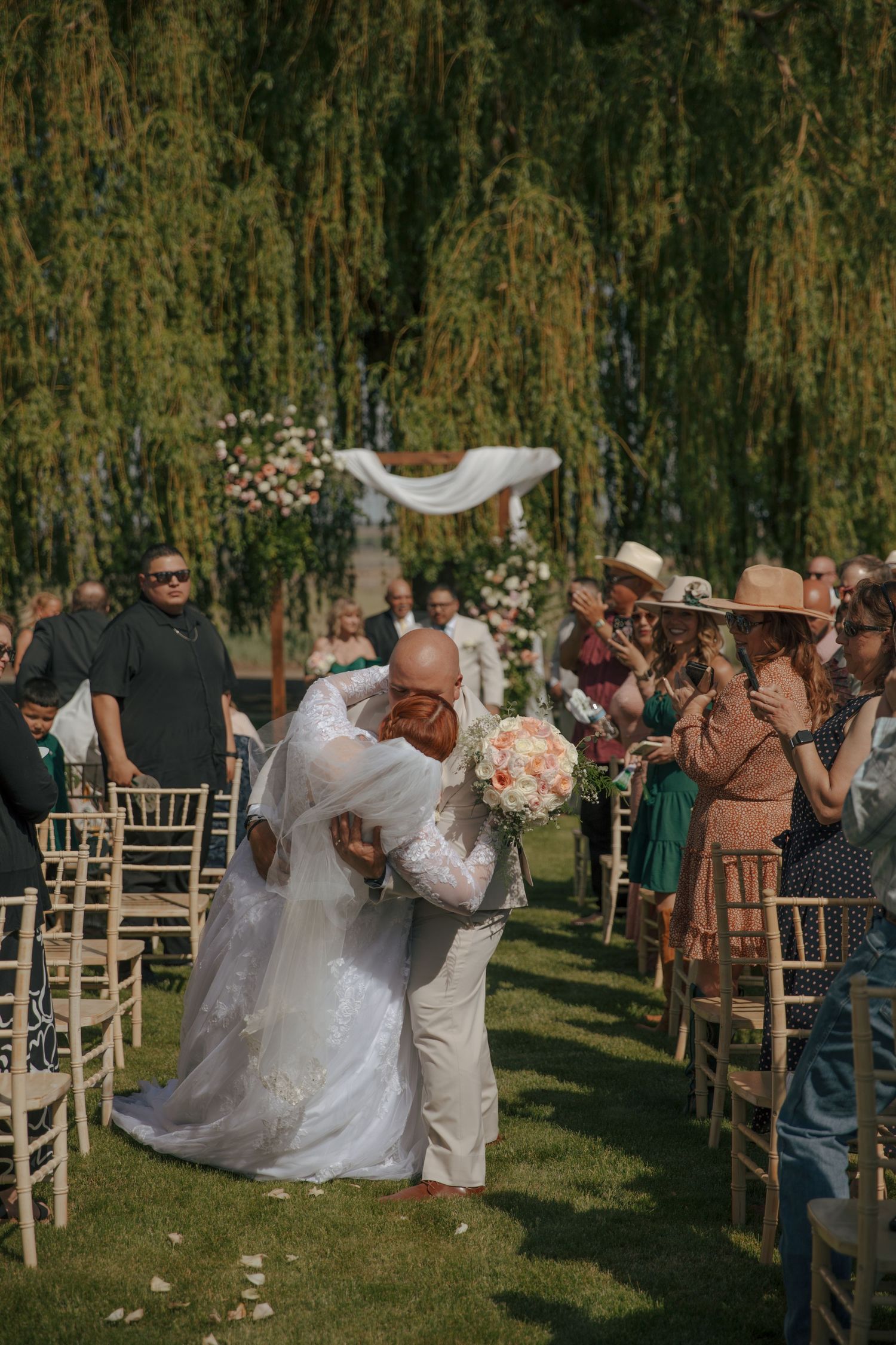 Bride and groom share a romantic dip kiss at outdoor wedding ceremony with guests applauding beside chiavari chairs.
