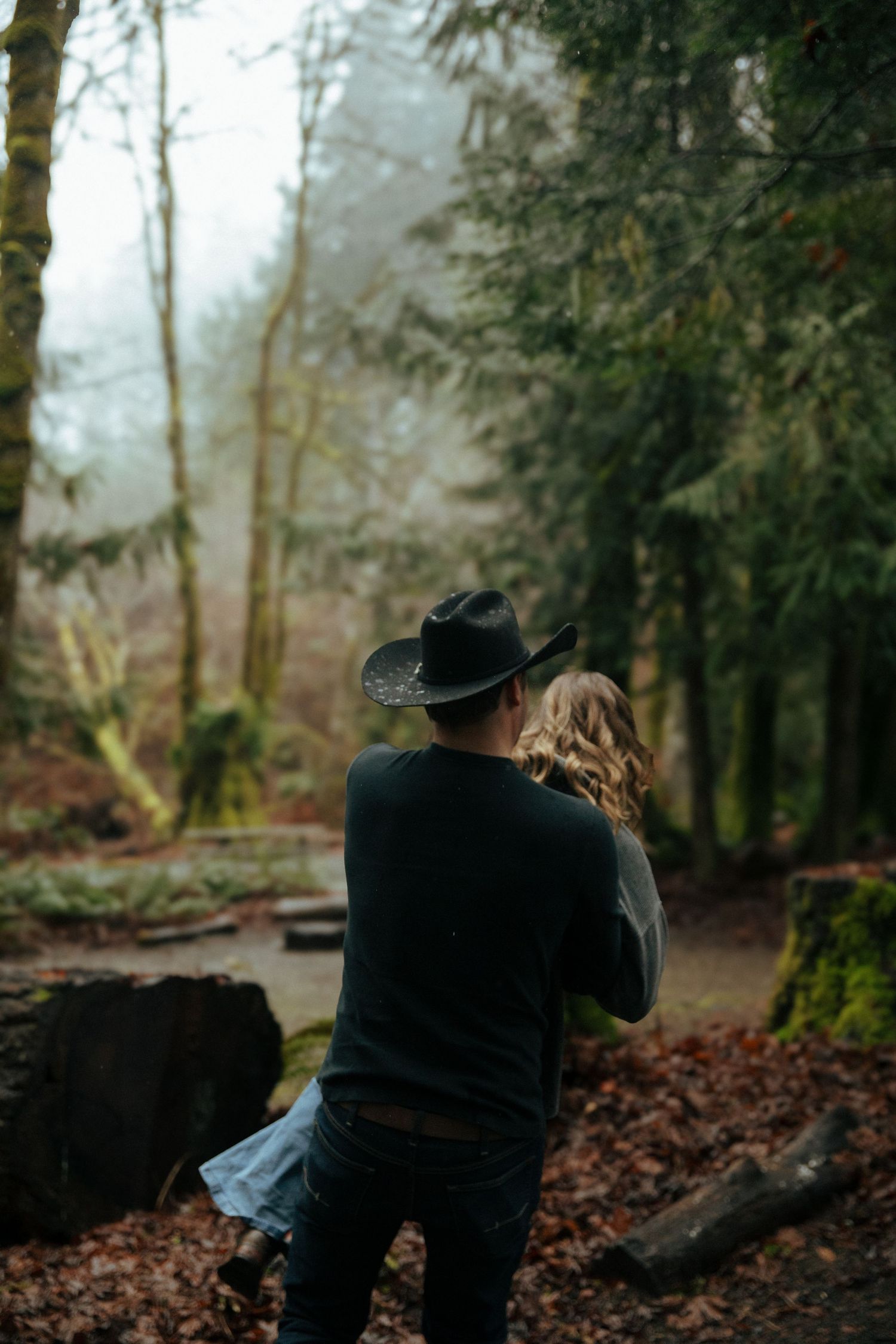 A couple embraces in a misty forest with fallen leaves and moss-covered logs creating a moody autumn atmosphere.