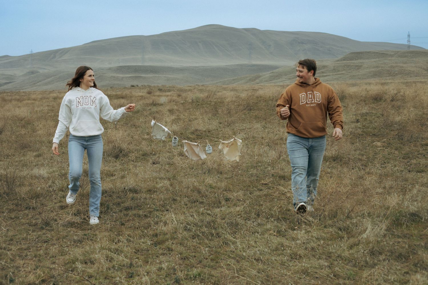 Two people walk through a grassy field with rolling brown hills in the background on an overcast day.