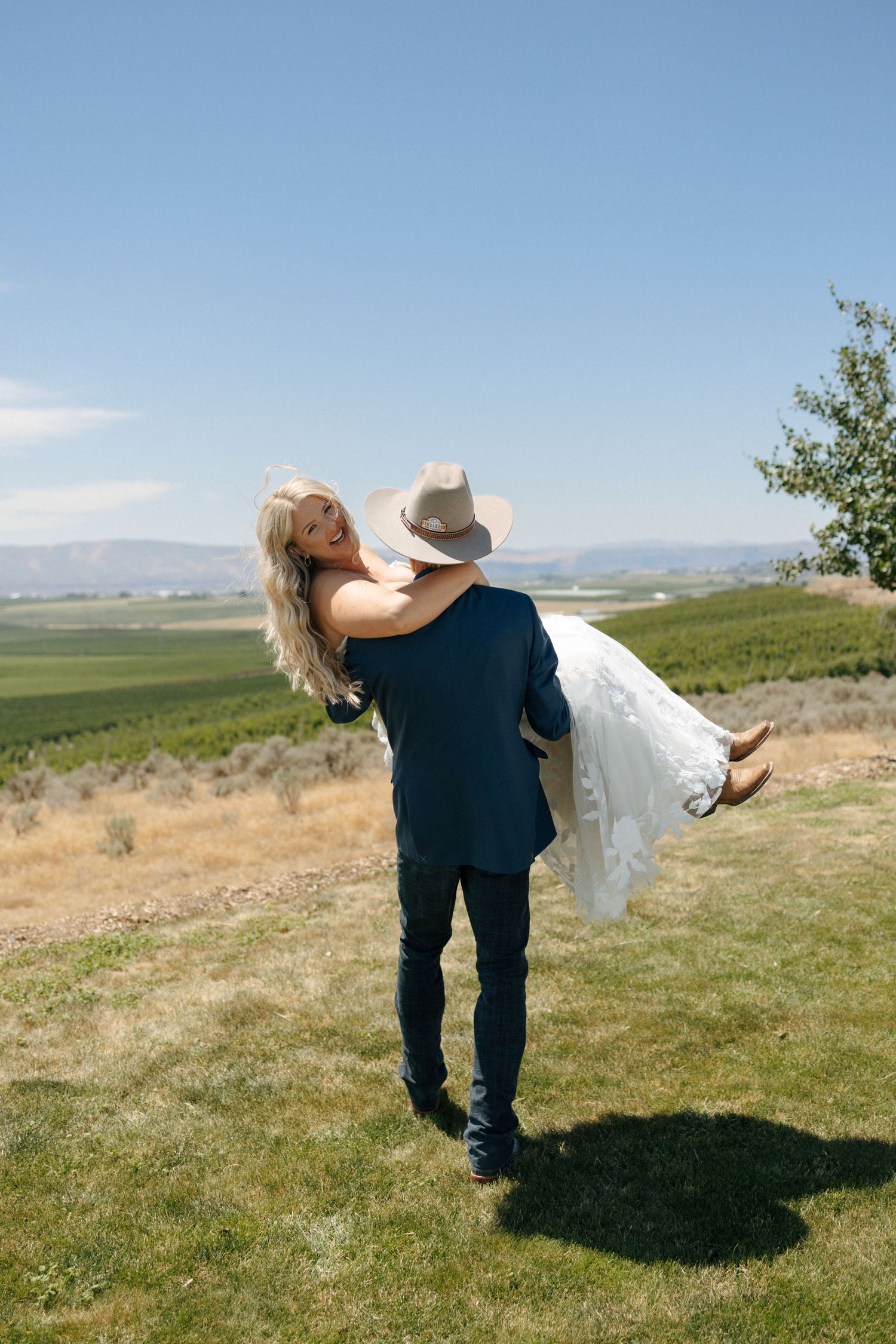 Groom in navy suit and cowboy hat lifts bride in white dress outdoors with scenic valley views.