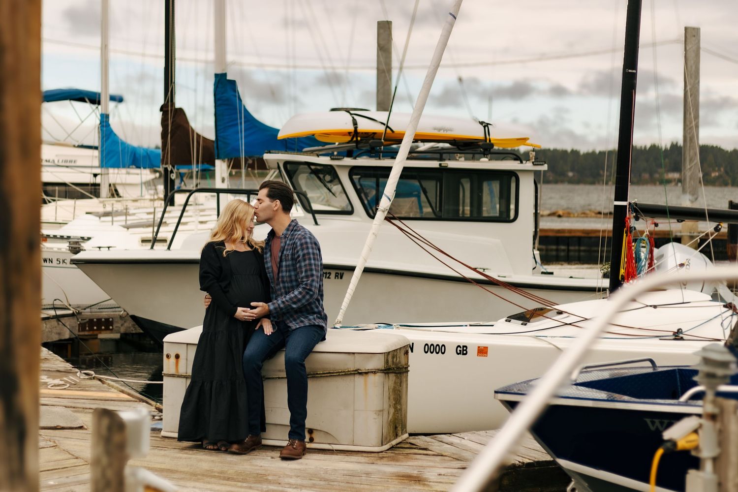 Couple sharing a romantic moment on a marina dock surrounded by sailboats on a cloudy day.