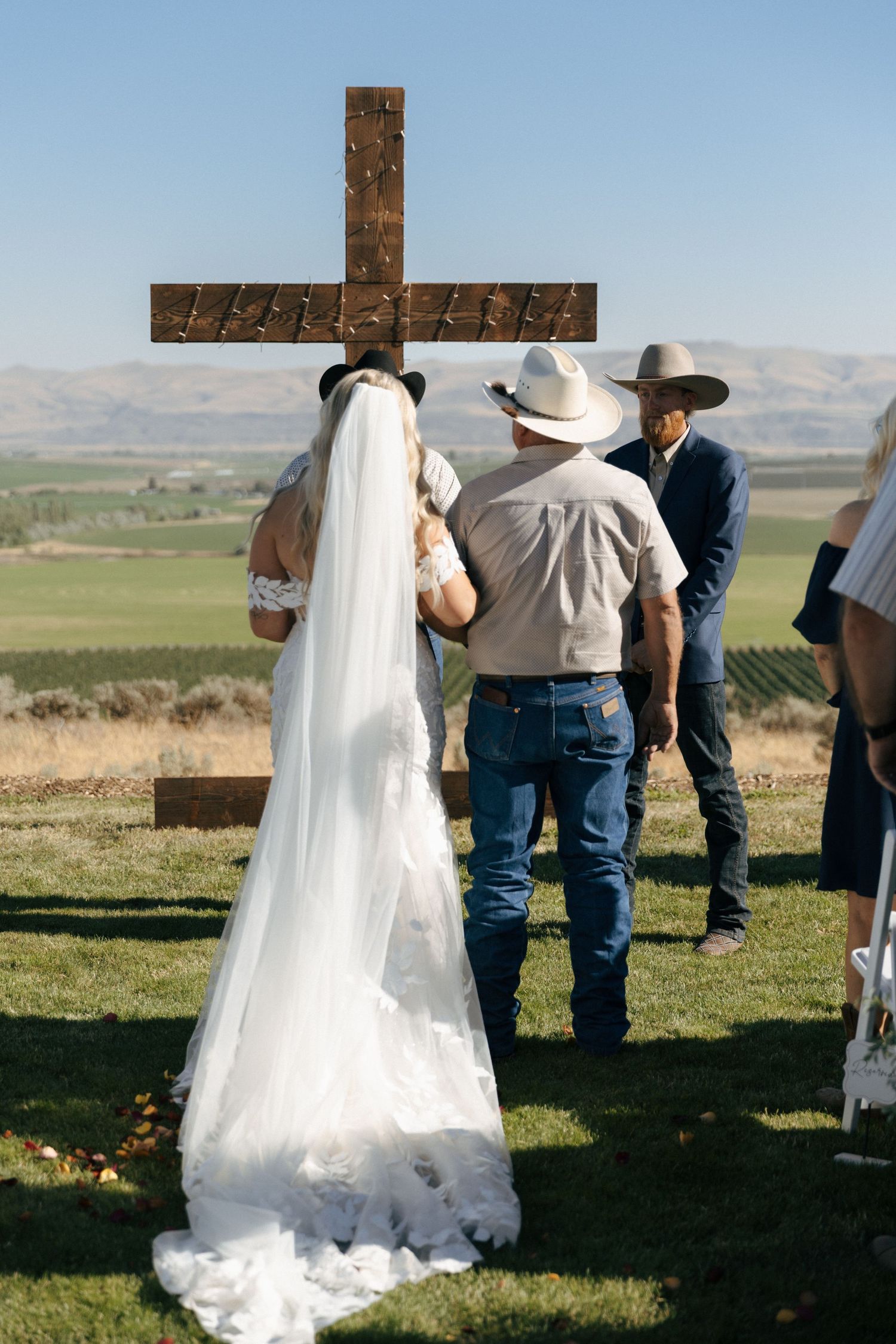 Outdoor wedding ceremony with bride in white veil before rustic wooden cross, cowboy-hat guest, scenic valley backdrop.