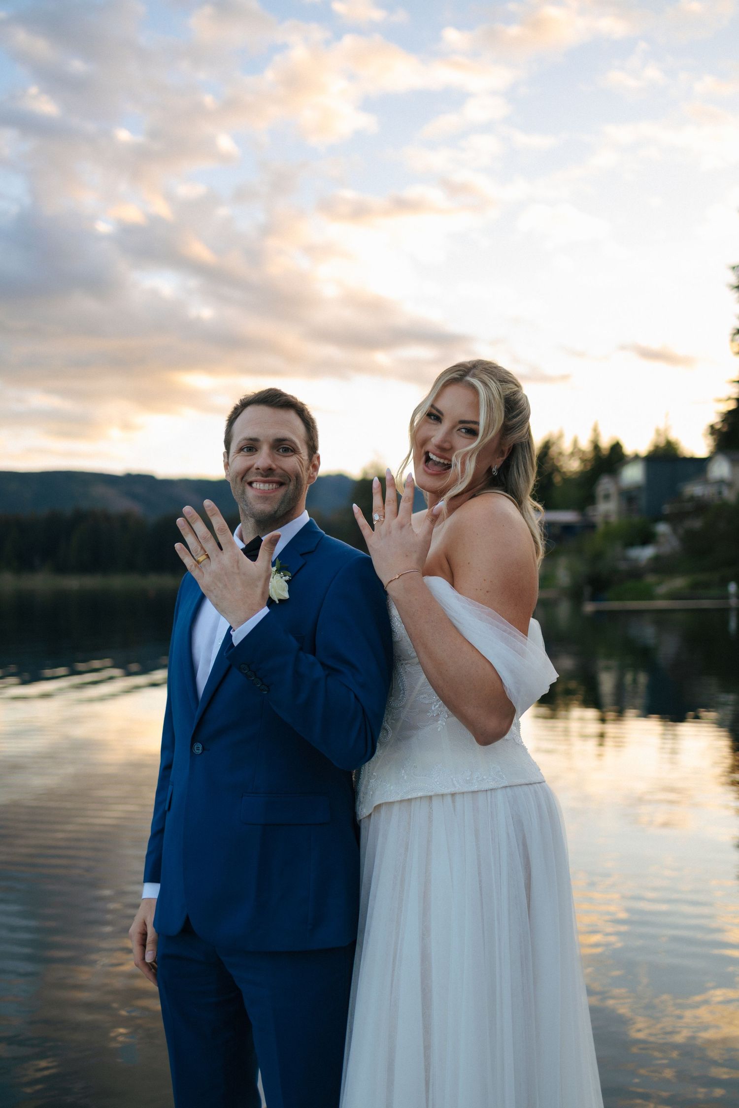 Newlywed couple showing wedding rings by a lake at sunset, groom in blue suit and bride in white gown smiling joyfully.