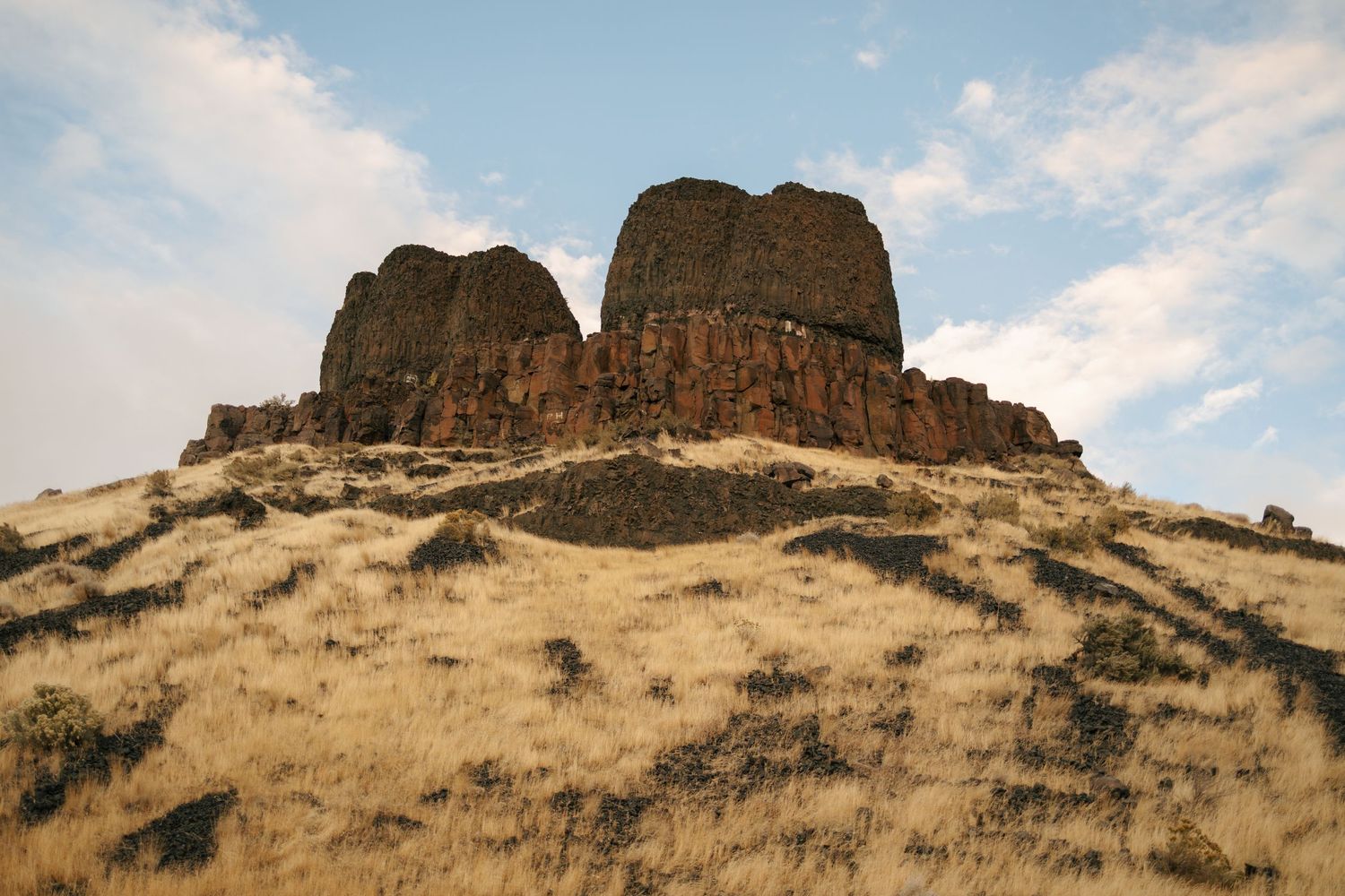 Large rock formations rise from a dry grassy hillside against a blue sky at Twin Sisters rock formation in Oregon.