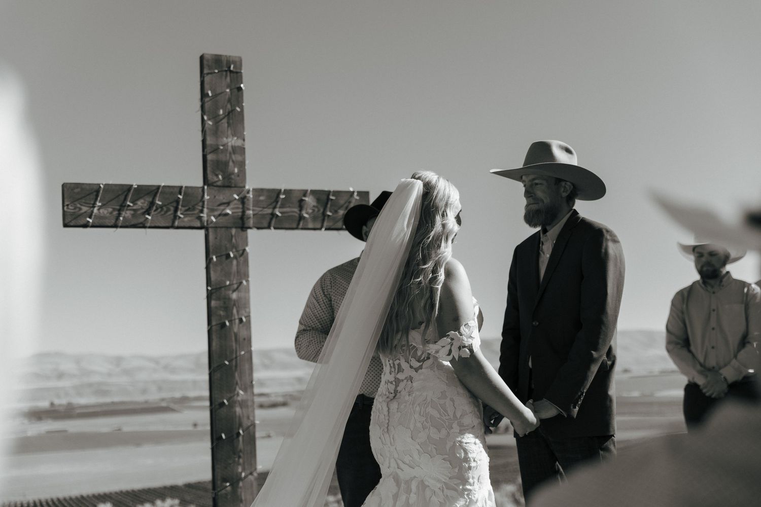 Black and white outdoor wedding ceremony with bride in lace gown and groom in cowboy hat kneeling before a rustic cross.