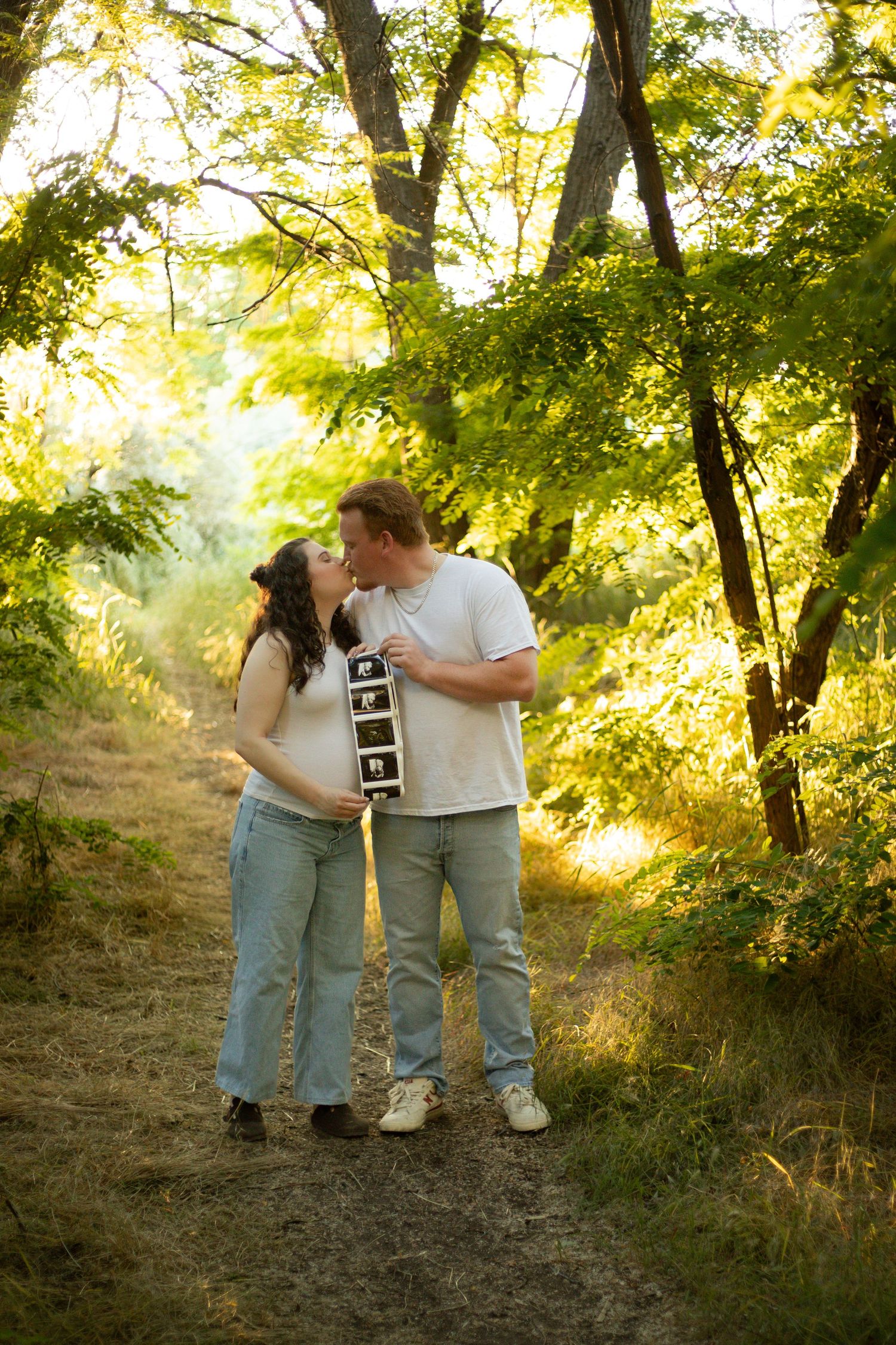 A couple shares an intimate moment on a sunlit forest path while holding ultrasound photos in their hands.