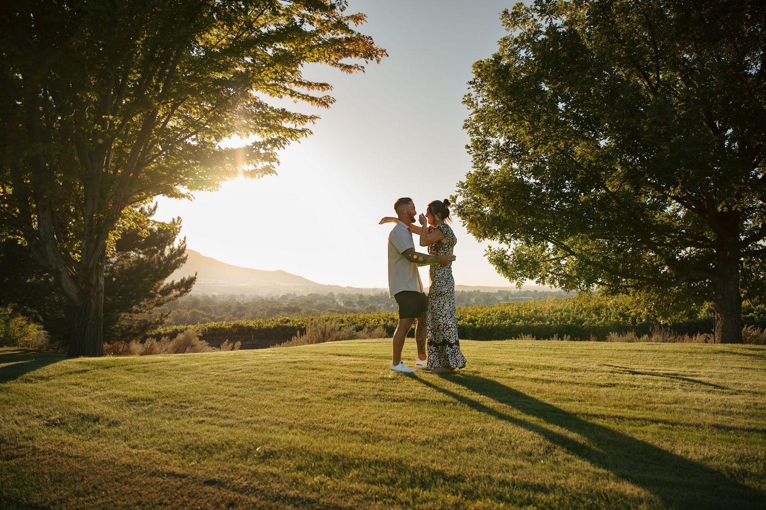 Couple dancing together on lush green lawn at golden sunset with trees and mountain views in background.